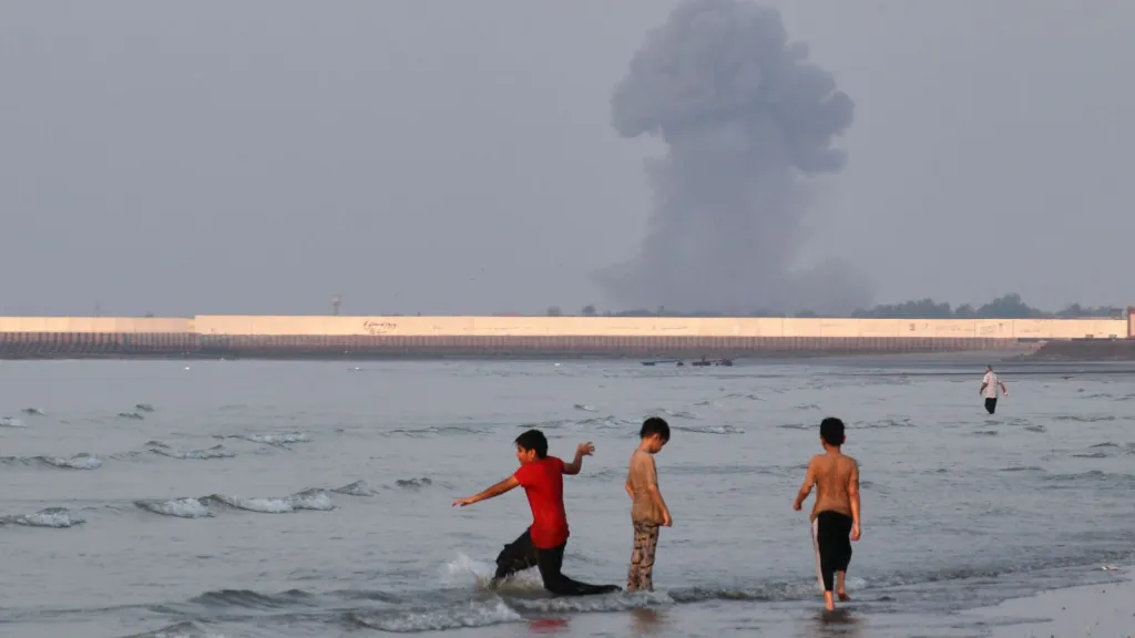Three boys play in the shallow waters of the Strait of Hormuz, as a plume of smoke rises from an explosion in the background, off Bandar Abbas, Iran. (Razieh Poudat/ISNA via AP)