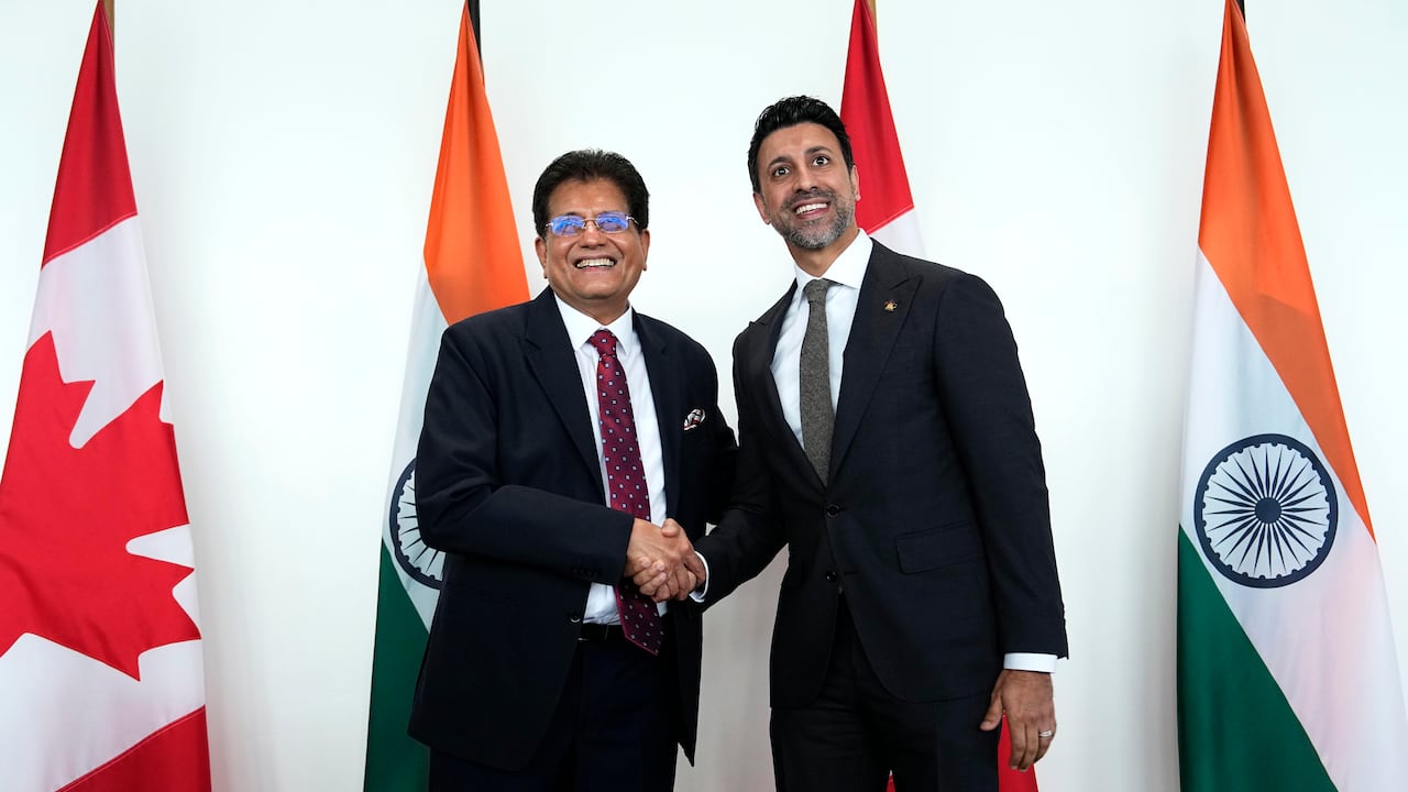 Two men with dark hair and dark suits smile as they shake hands.