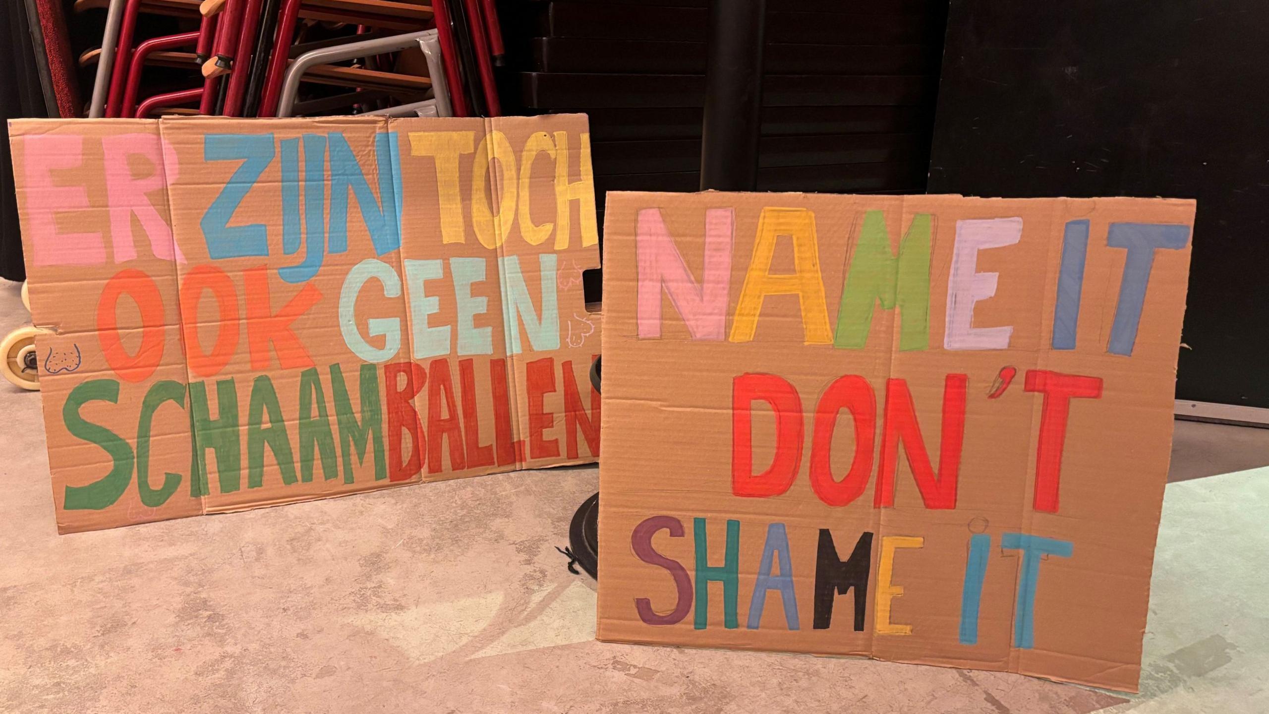 A photo of two pieces of cardboard with large writing in multiple colours. One is in Dutch and translates as "afterall there are no shame balls" and the other says "name it don't shame it". 