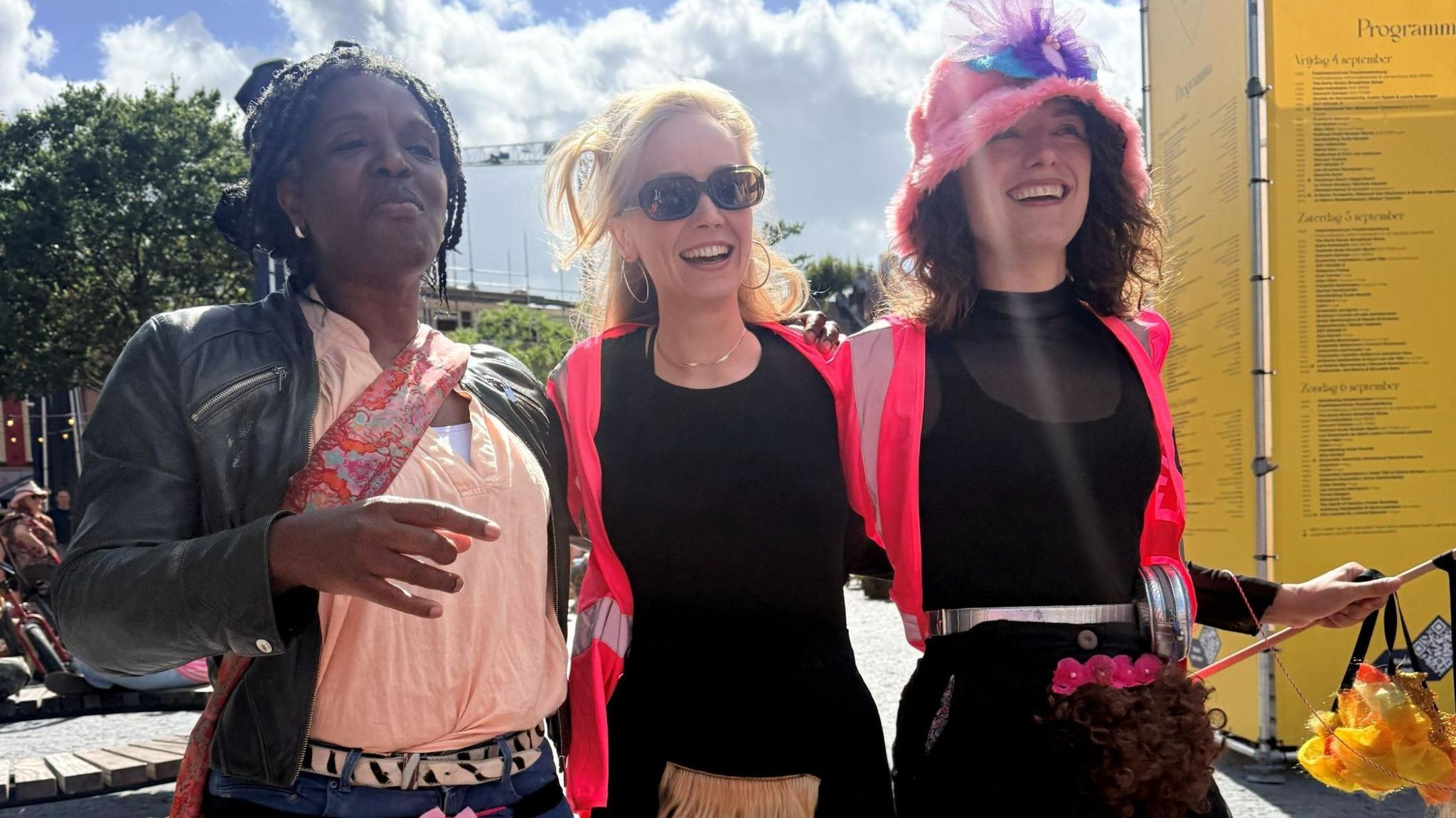 Three people who joined in the march.  Two wear bright pink jackets, the other a pale pink shirt. They are smiling and wearing merkins - artificial pubic hair pieces - which sits over their clothing. 