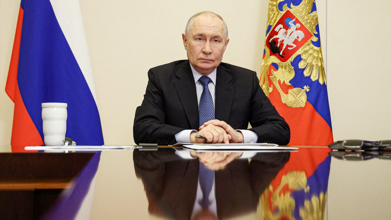 A man sits at a table, mirrored in its polished surface and flanked by Russian flags