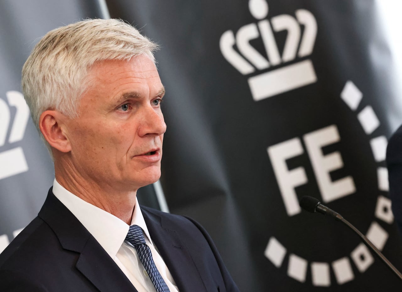 A man in a suit stands next to a black flag bearing the letters 'FE'