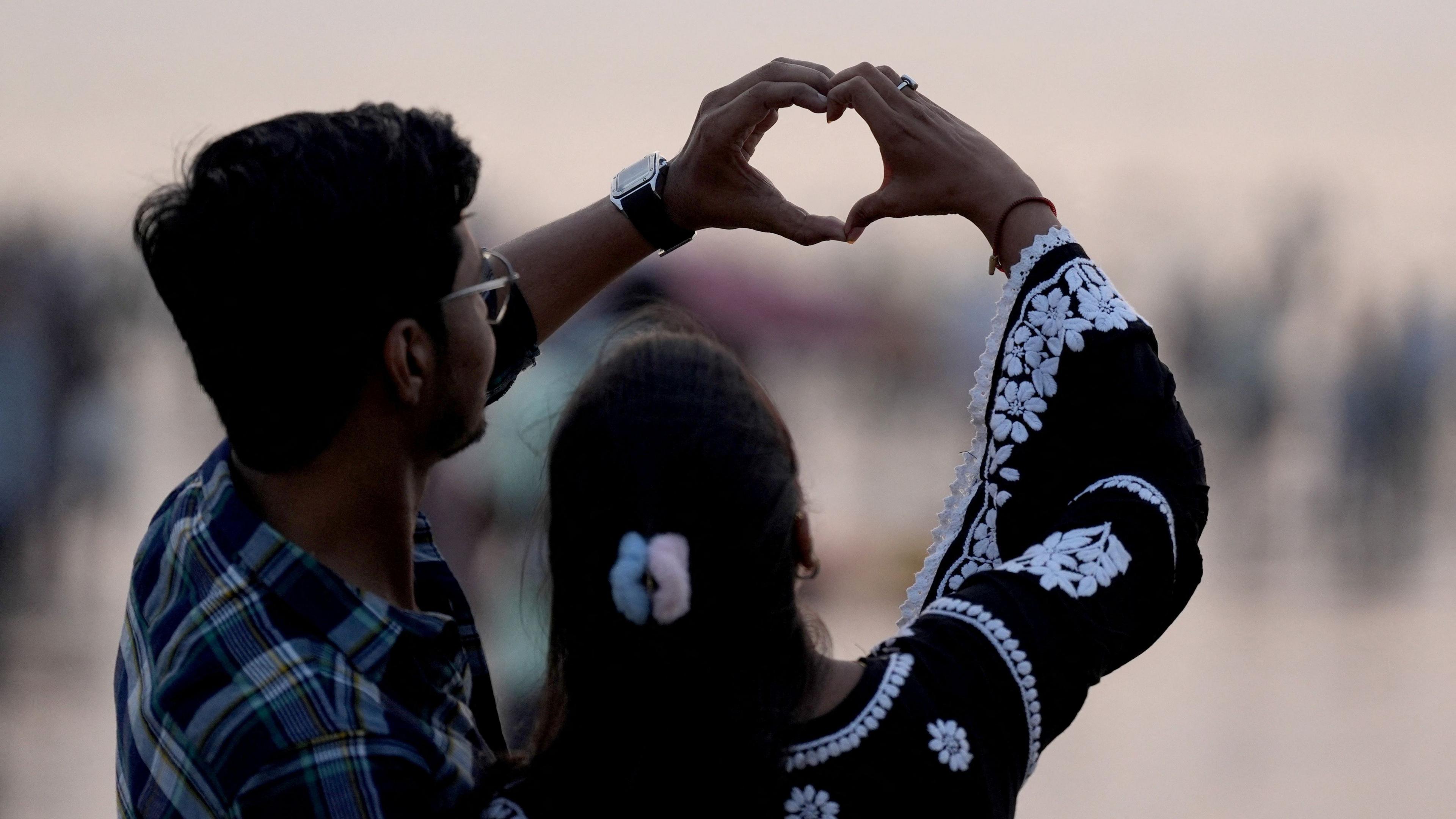 A backshot of a couple - with the man wearing a check shirt and the woman dressed in black and white tunic - holding their hands up against the Mumbai skyline to make a heart