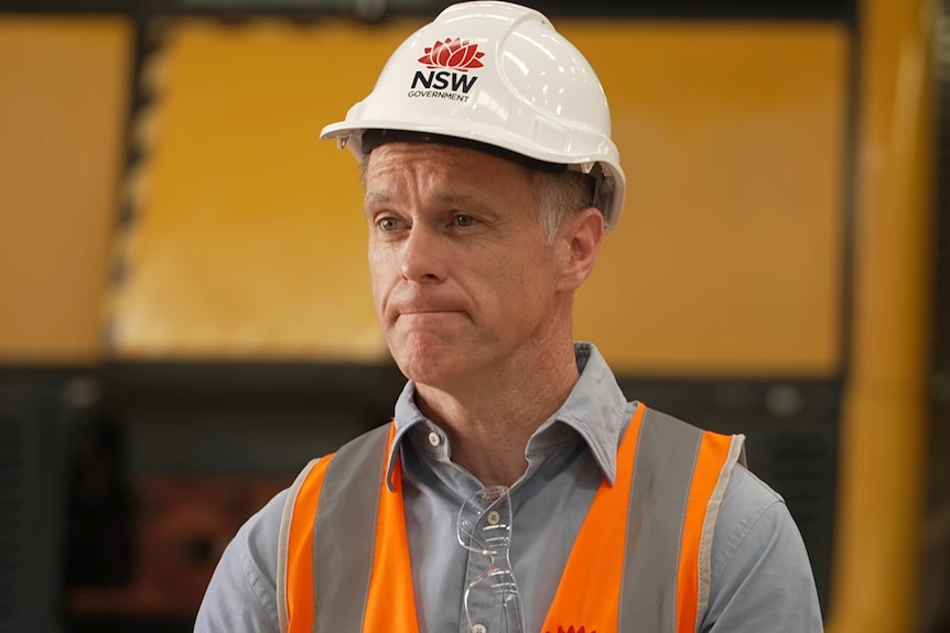Chris wears a hard hat and high-vis vest. His lips are pressed in a stern expression.