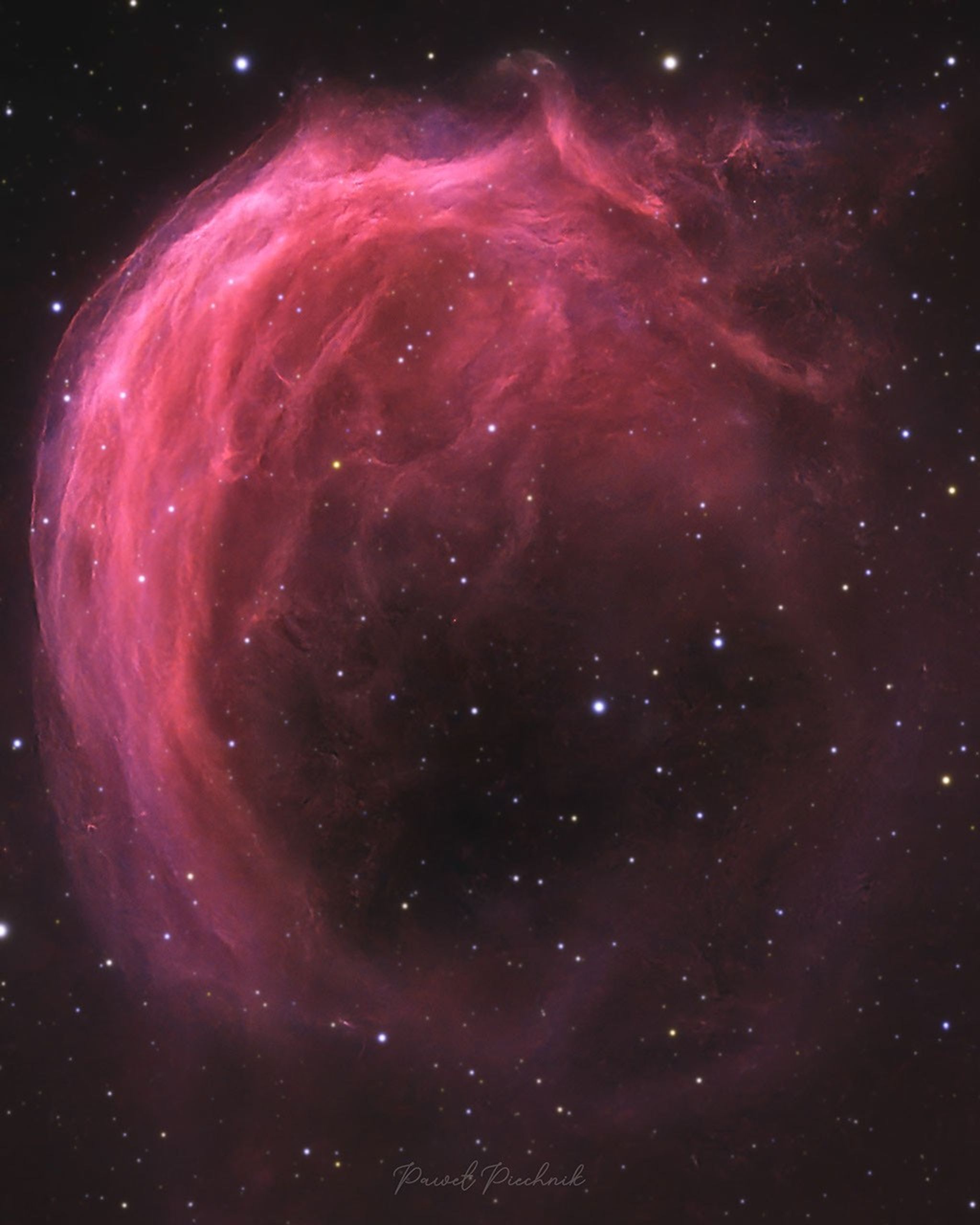 A starfield has a bright pink swirling nebula  in it. The top part of the nebula shows much detail, while the bottom part is mostly open.