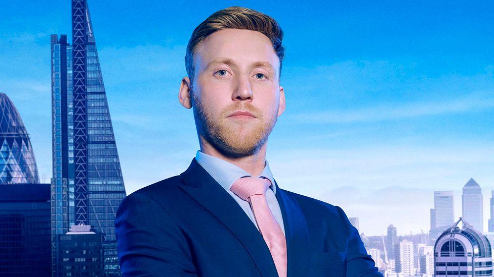 Studio publicity portrait of a person in a navy suit, light blue shirt and pink tie standing with folded arms against a London skyline backdrop featuring The Shard and Canary Wharf buildings.