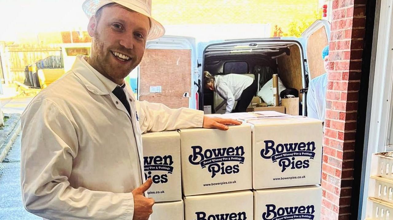 A man with a cropped brown beard is wearing white overalls and a white hat. He is smiling and has a thumbs up by a stack of white boxes that have 'BOWEN PIES' written on them in blue. 