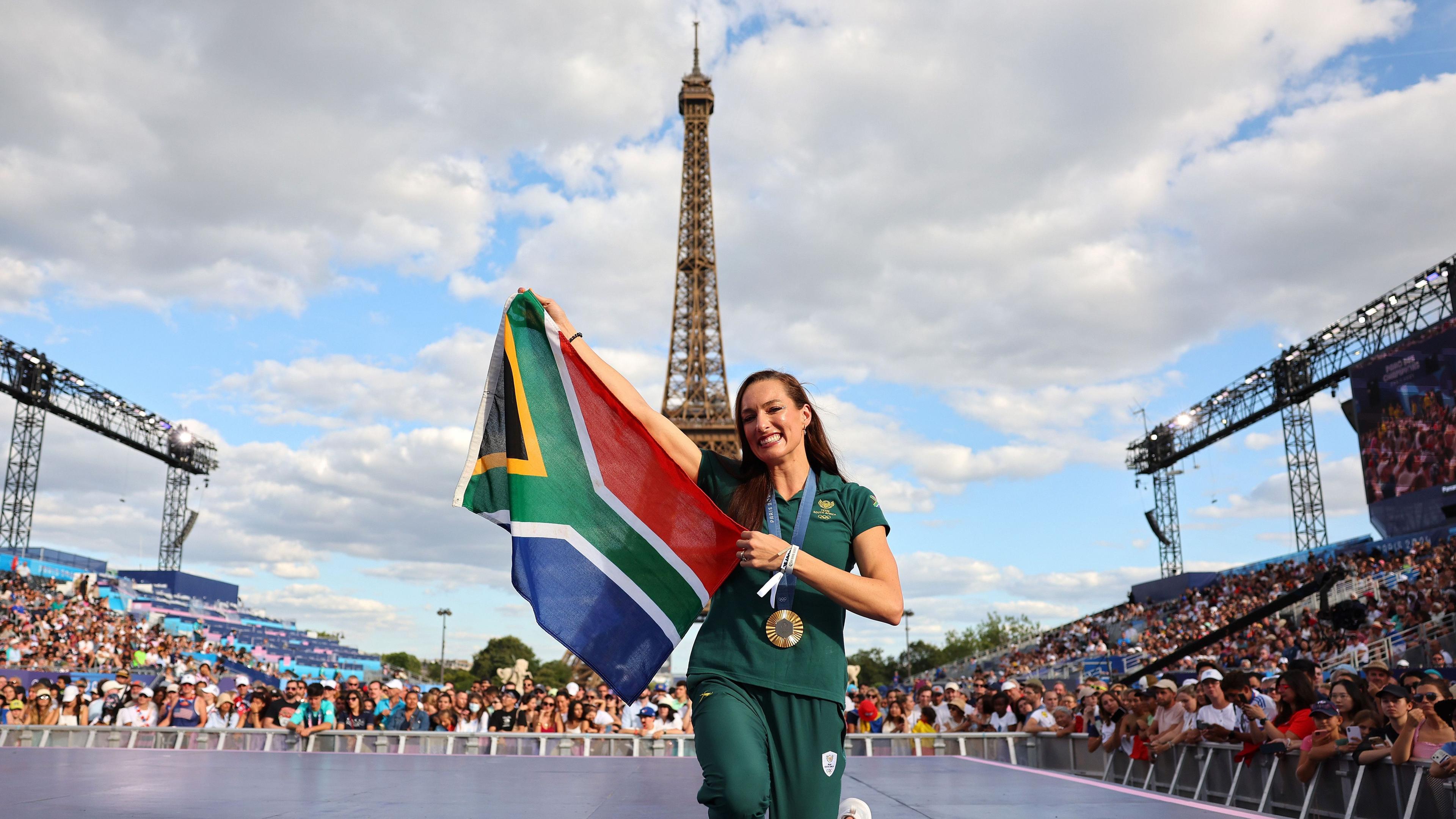 Tatjana Smith, wearing dark green South Africa tracksuit bottoms and shortsleeved training top, smiles as she kneels on a large raised stage and holds up a South African flag to her right. The swimmer also has an Olympic gold medal on a ribbon around her neck. A large crowd of people can be seen gathered around the stage, with some leaning over barriers at the front and others seated in gently sloping stands, while in the background the Eiffel Tower rises directly behind Smith into a blue sky do