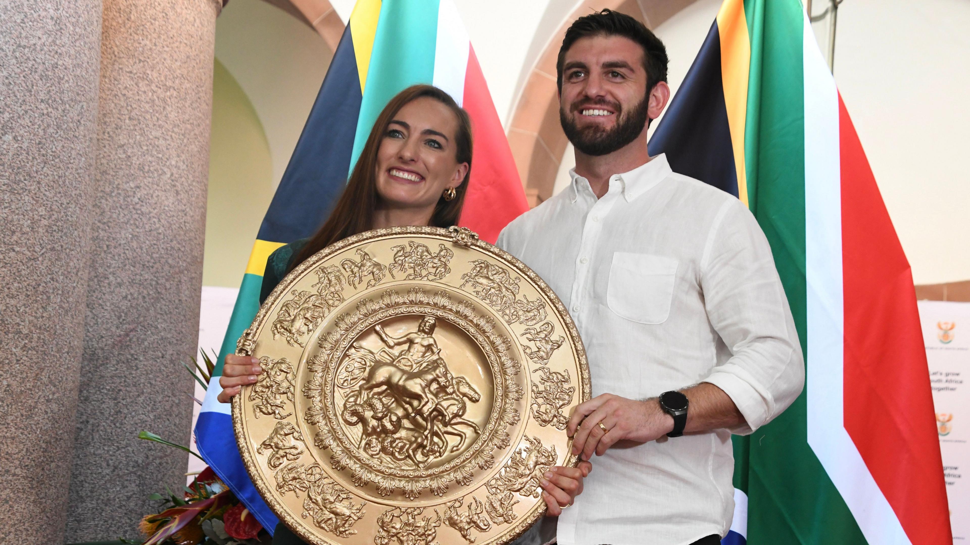 Tatjana Smith and her husband Joel Smith, visible from waist up, stand side-by-side as they hold up a large and ornate circular gold trophy. The centre of the embossed plate appears to show a man riding a chariot driven by more than one horse, with the outer circumference also covered in classical-looking designs. Behind the smiling couple hang two South African flags.