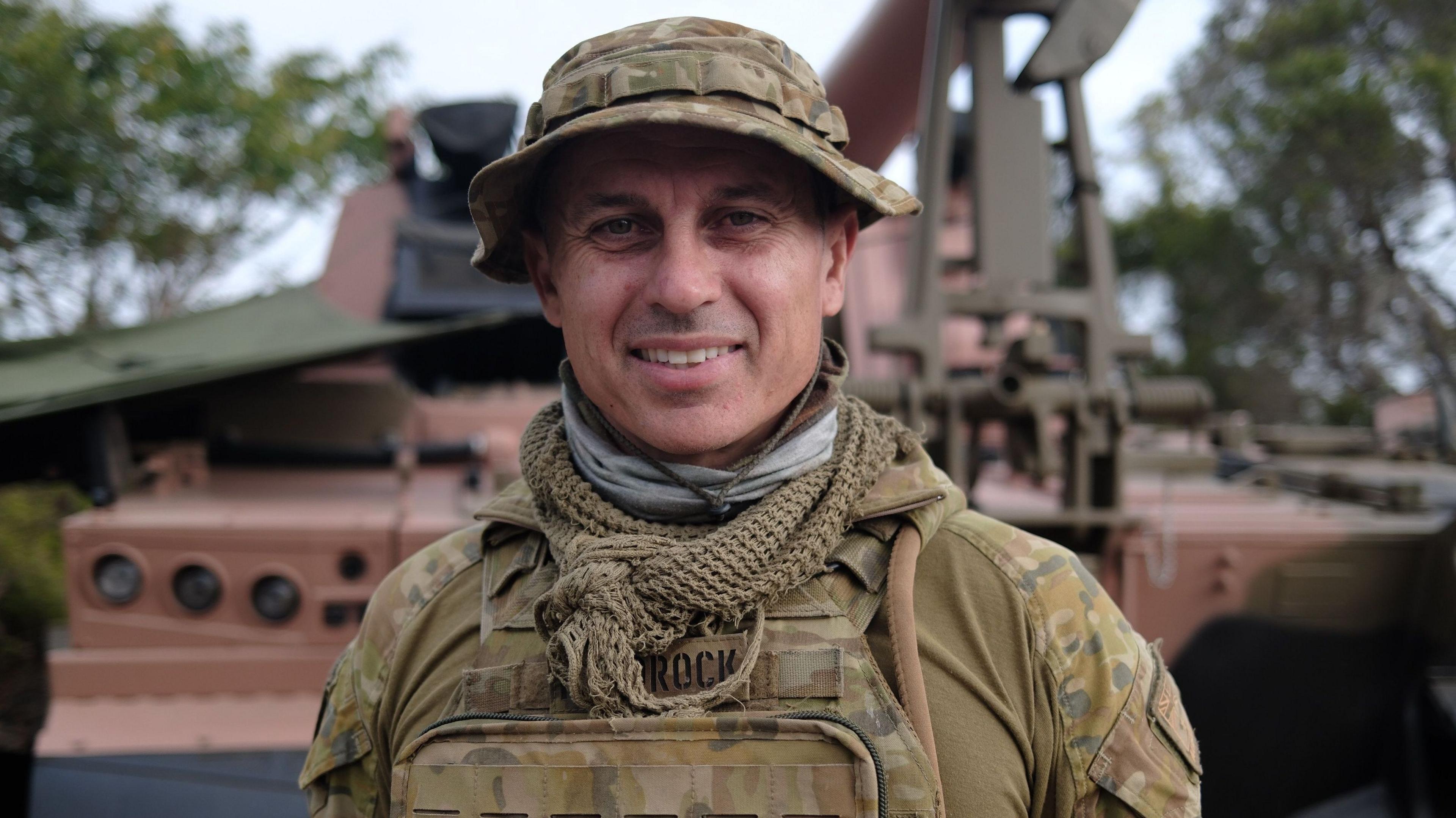 A man in army fatigues smiles at the camera with a tank behind him
