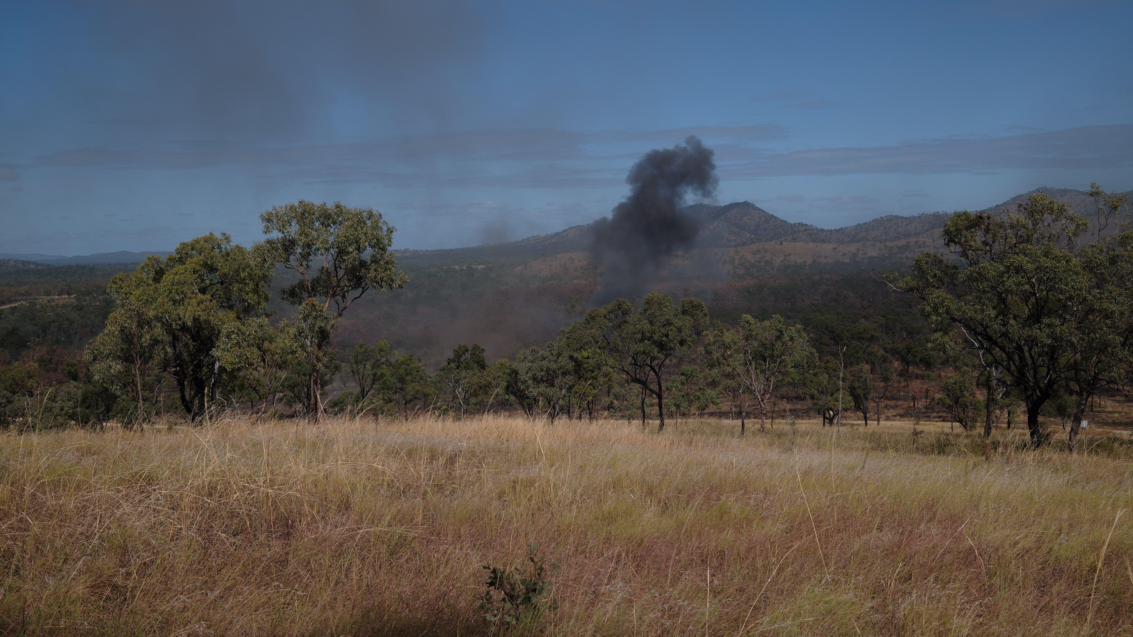 A picture of dry grassland with trees in the middle distance and black smoke rising with hills in the background