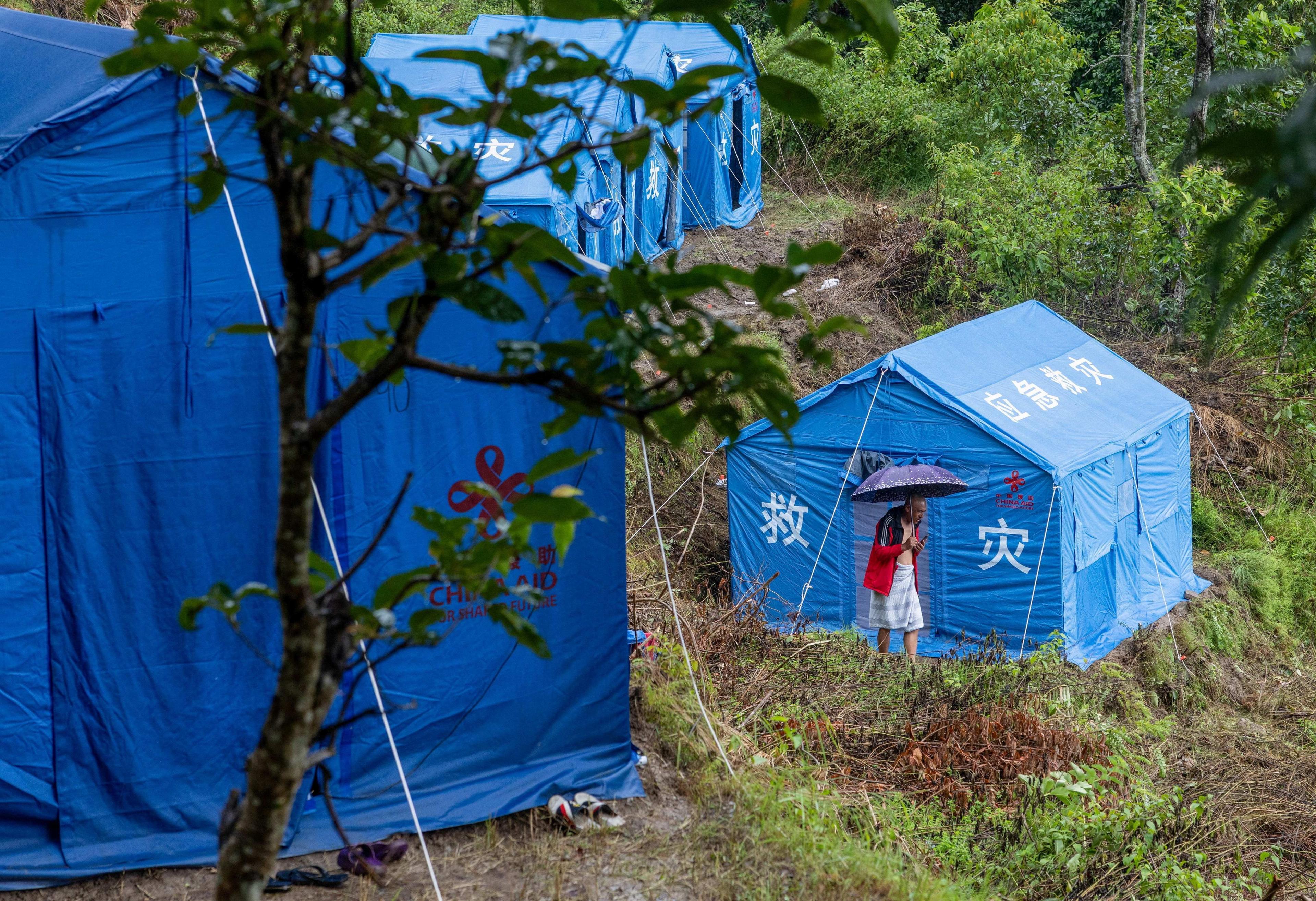 Blue disaster-relief tents on a muddy, forested hillside. A person holding an umbrella stands outside one of the tents. White Chinese characters and a relief-aid logo are visible on the bright blue tent walls.