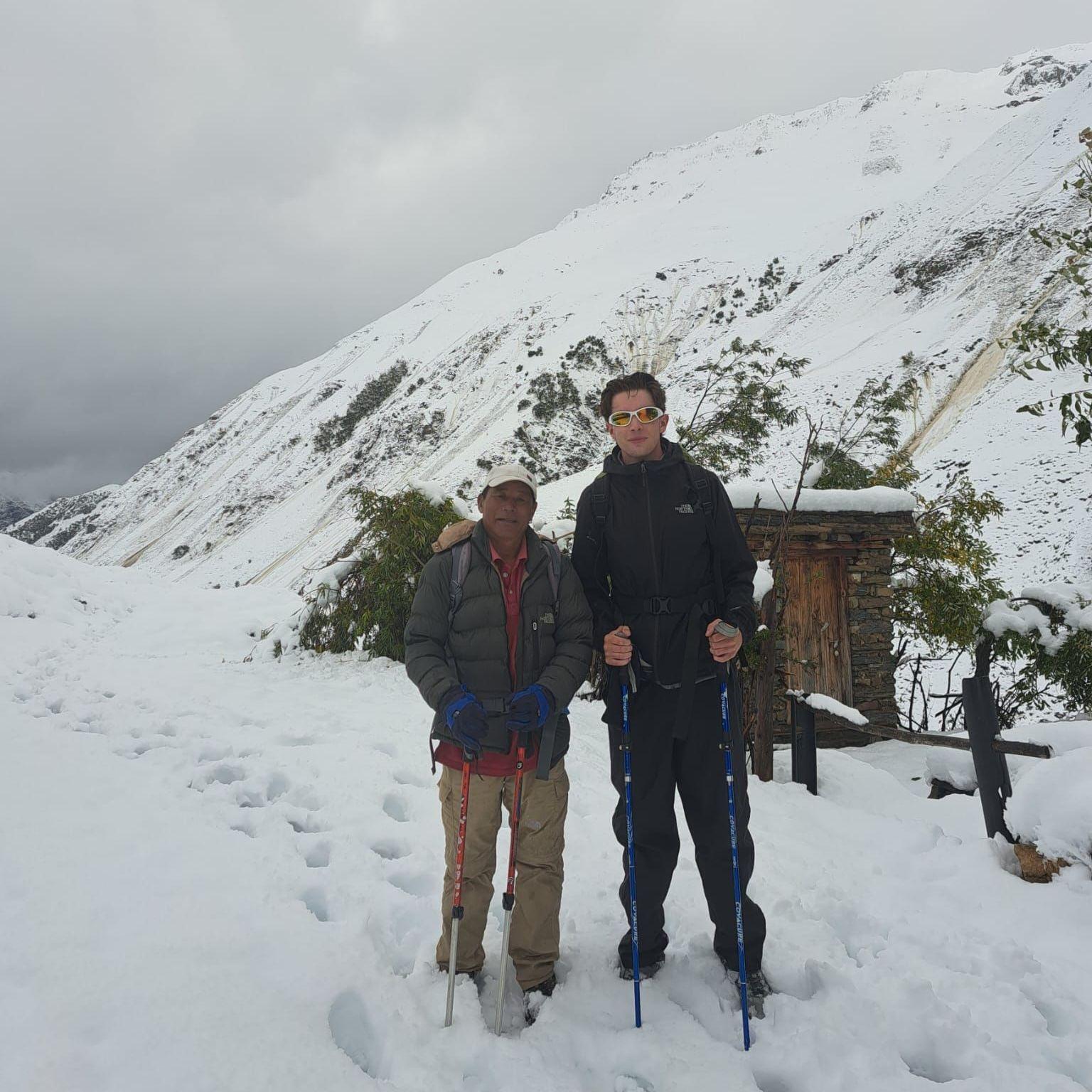 Jake Stacy and his Nepalese guide stand on a snow-covered mountain trail holding trekking poles, with steep white slopes rising behind them. A small stone hut partly covered in snow sits to one side of the path, while low clouds and an overcast sky surround the snowy landscape