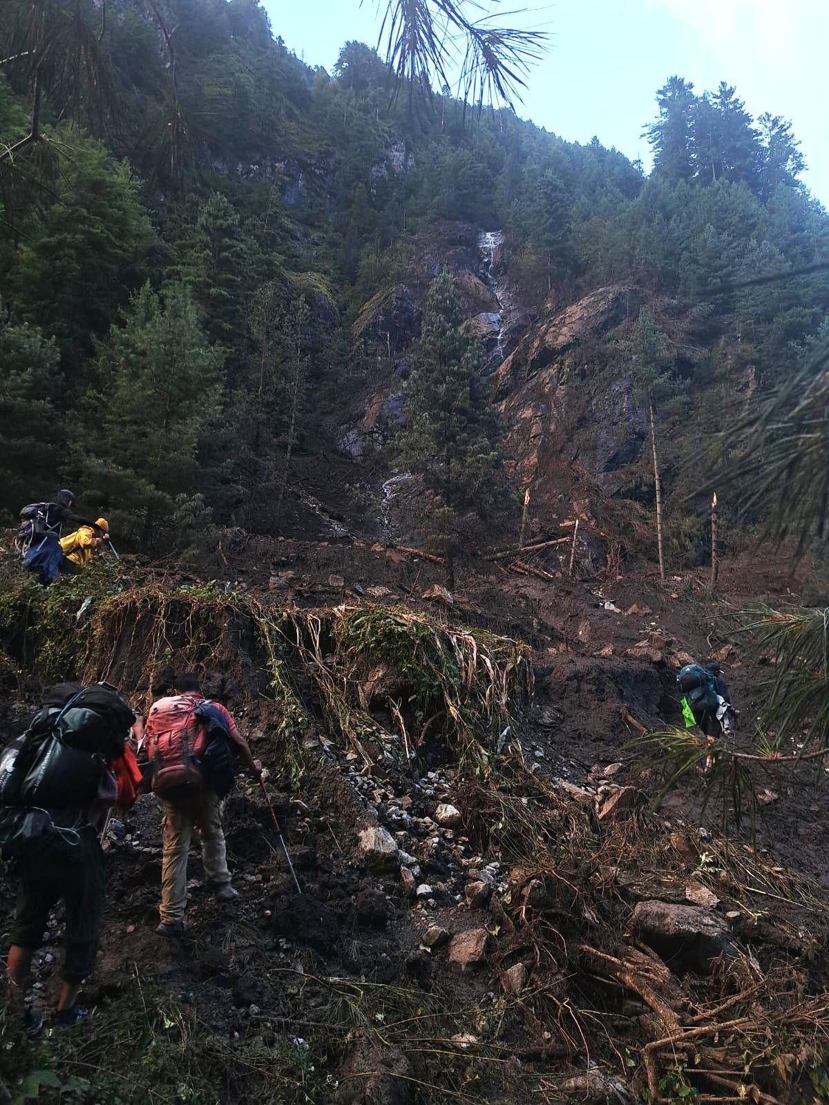 Hikers carrying large backpacks and trekking poles climb across a muddy, debris-strewn slope in a dense mountain forest. The trail appears damaged by a landslide, with exposed earth, rocks and uprooted vegetation.