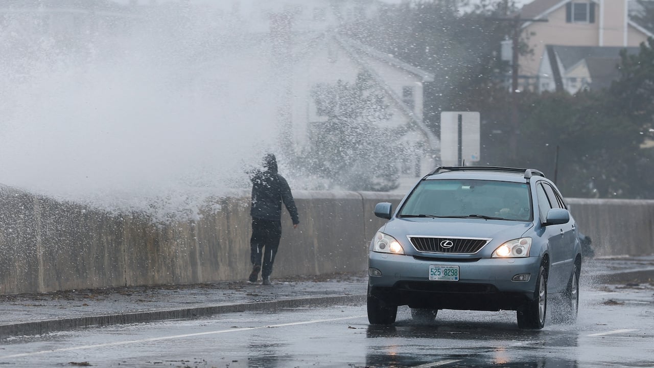 A wave crashes against a seawall as pedestrians and cars navigate a street.