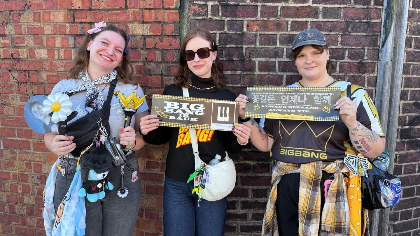 Three women standing in front of a brick wall display concert merchandise and banners. One  holds a flower-shaped light stick and a crown-shaped light stick, with a cross-body bag, keyrings and a blue shopping bag. The centre woman holds a banner reading “BIG BANG IS BACK” and carries a white shoulder bag. The third holds a banner with Korean and English text and wears a clear waist bag. Various accessories, badges and merchandise are attached to bags and clothing.