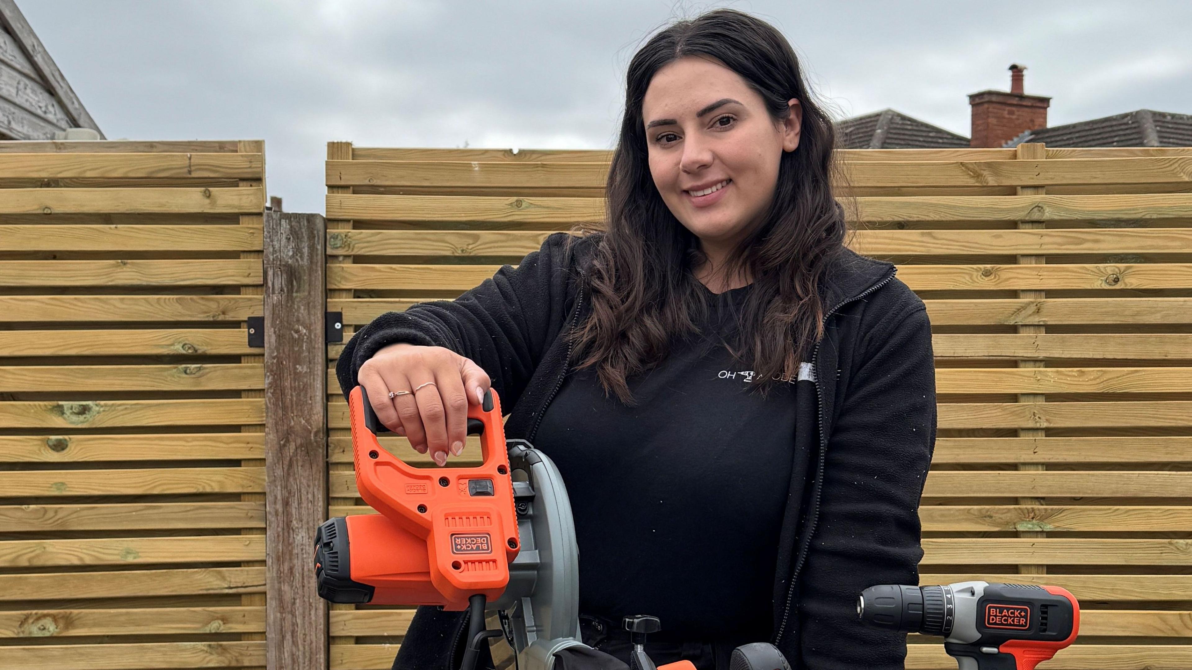 DIY influencer Jasmine Gurney stands outside in front of a fence, holding an orange and black power tool.
