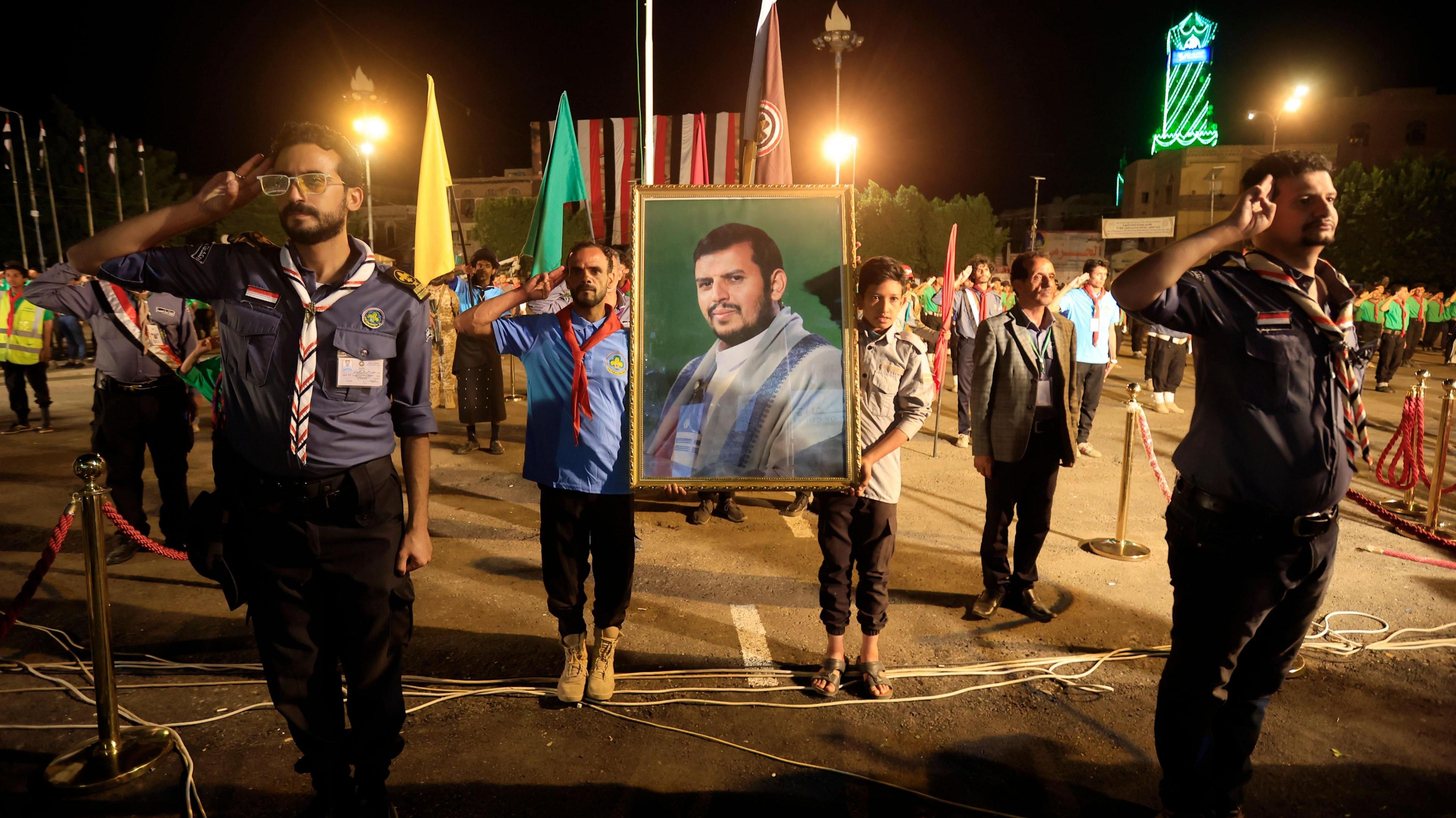 Houthi scouts celebrate the 12th anniversary of the takeover of northern Yemen in Sanaa. Several men in scout uniforms stand to attention and salute in an evening ceremony. Two in the centre hold a portrait of Houthi leader Abdul Malik al-Houthi