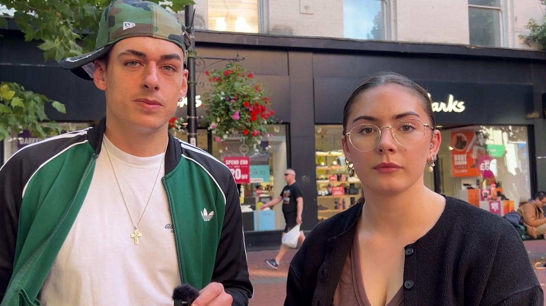 Cara, a young woman, who wears thin glasses and a black cardigan, and Dylan, a young man wearing a camo baseball cap sideways and a black and green jacket over a white t shirt