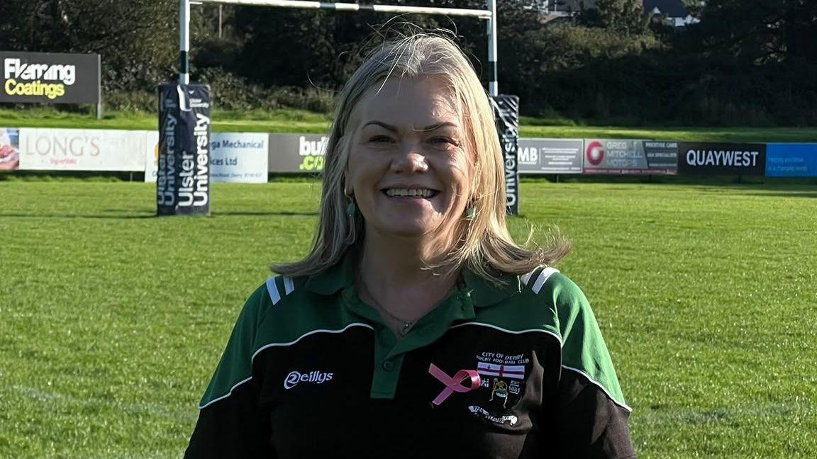 Women with blonde hair staring at the camera. She is wearing a green jump that says City of Derry women's rugby club.
