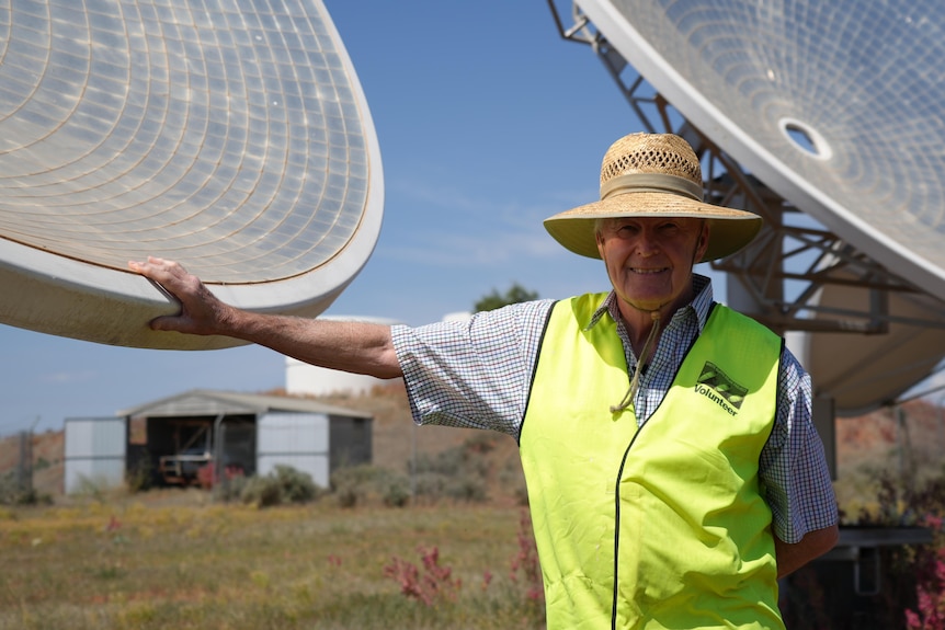 Graeme Hanigan wearing a hi vis vest and a sunhat, leaning on one of the parabolic dishes at the solar power station