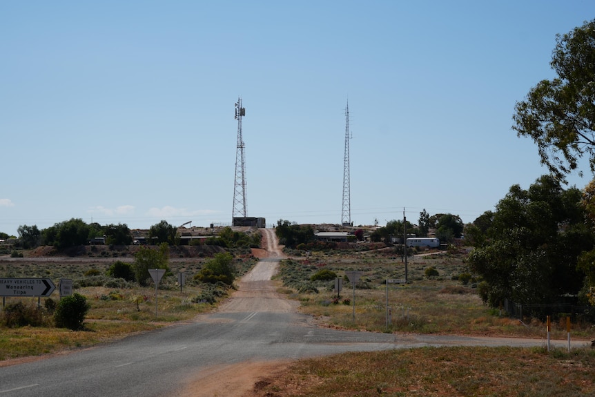 A bitumen road leading slightly upwards towards a small hill with houses and two satellite towers, under a clear blue sky 