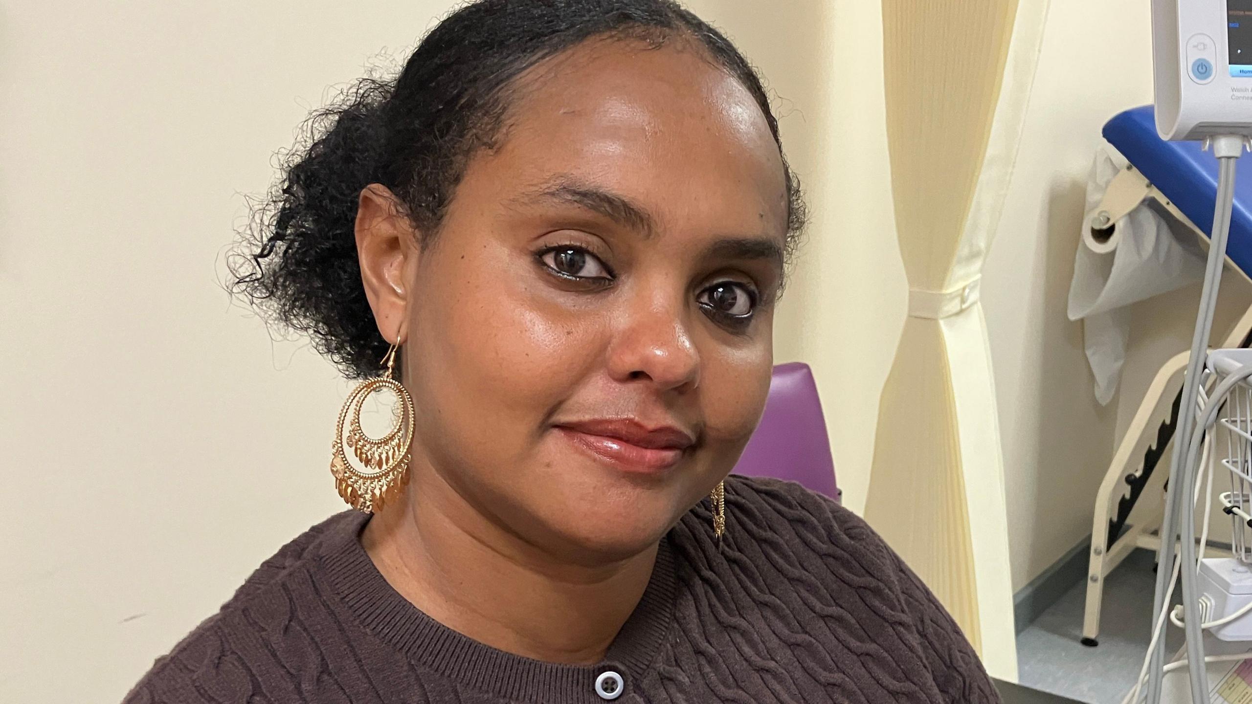 A woman with black hair, tied back, large gold earrings is smiling at the camera.  She is wearing a brown cardigan.