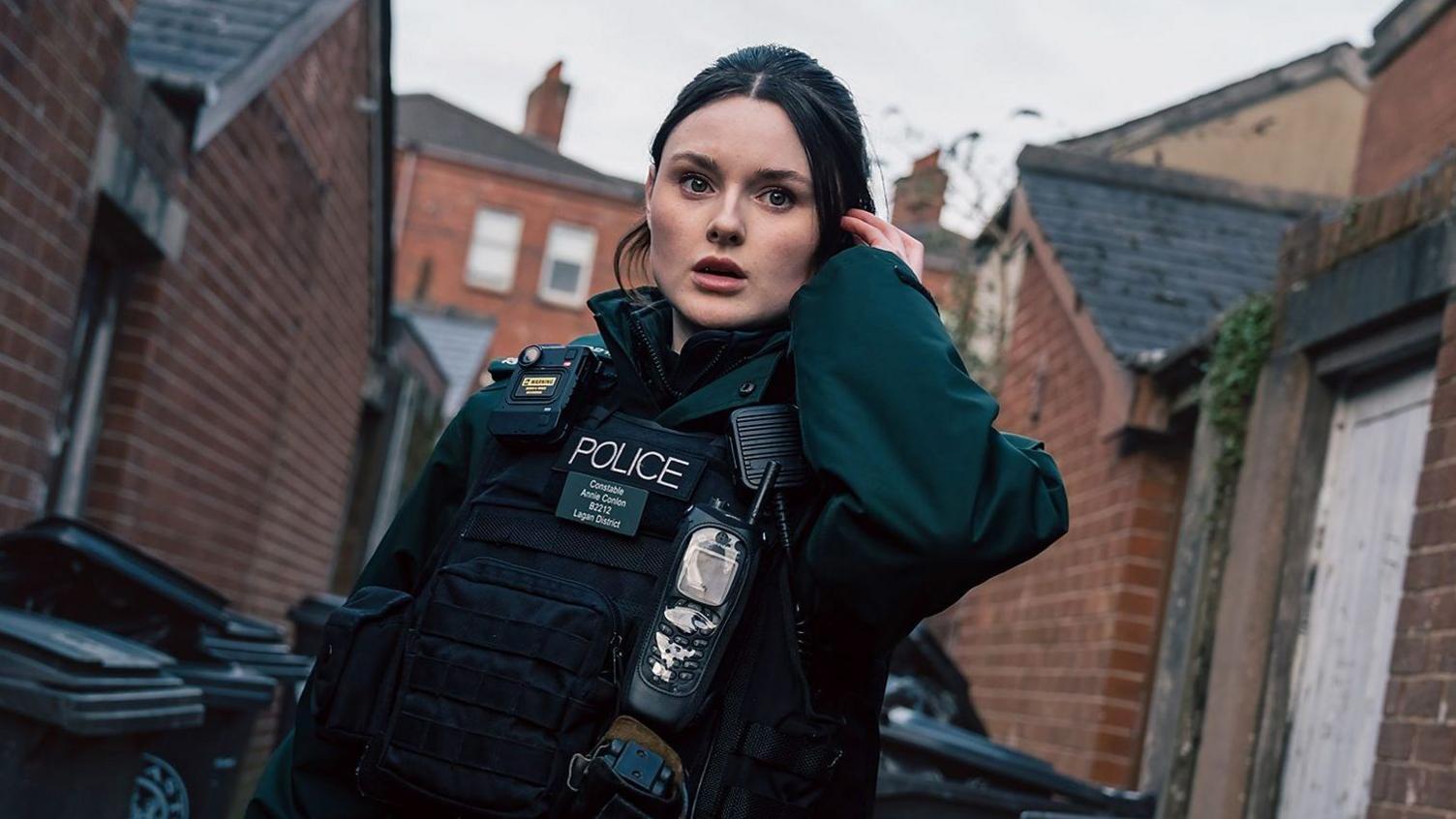 A young woman with brunette hair dressed in a dark green police uniform which includes a body-worn camera, stab vest and walkie-talkie. She's standing in an alleyway with red brick houses and bins alongside her. She's holding an ear piece to her left ear. She has a stunned impression on her face. 