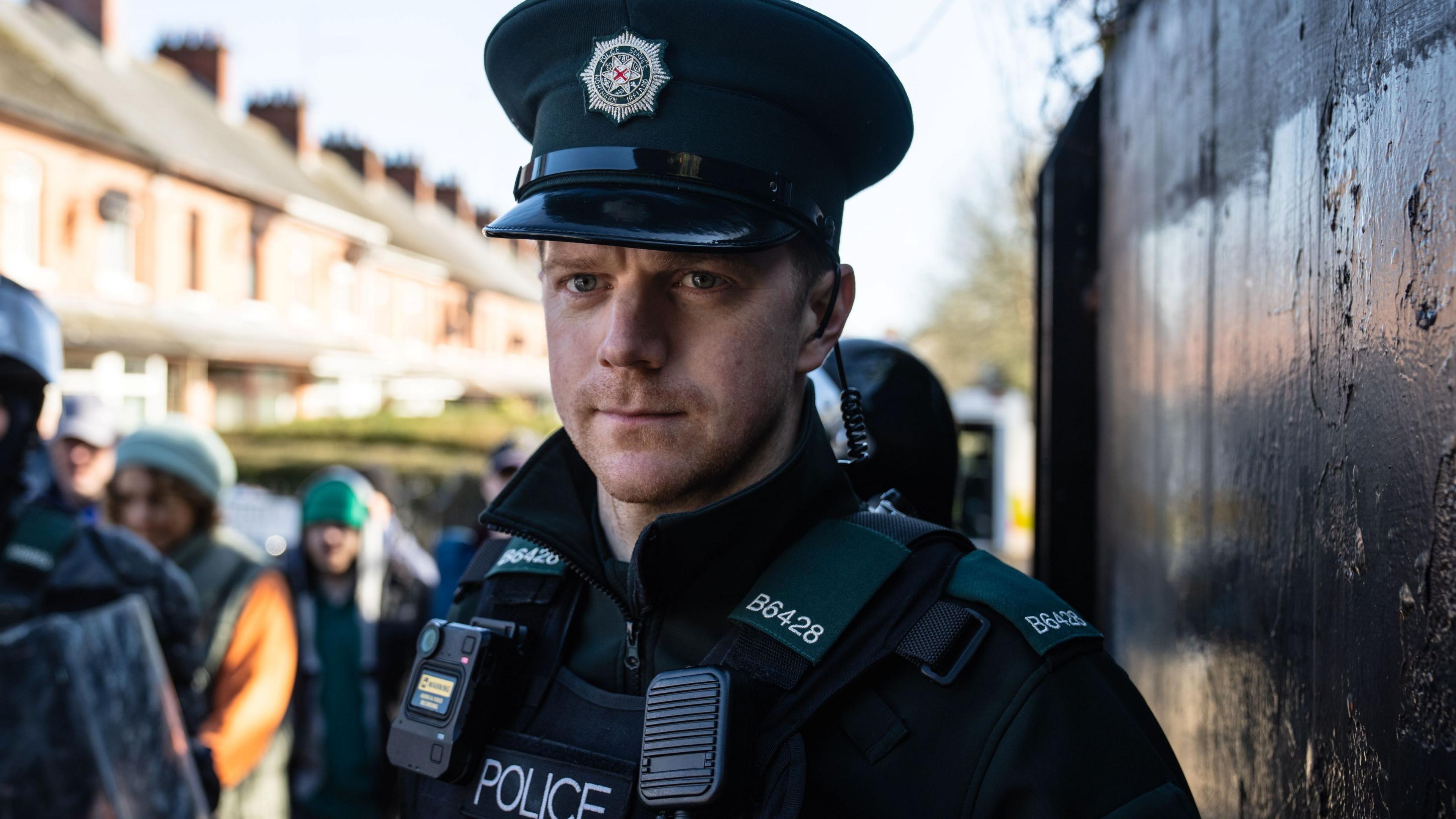 Shane is wearing a dark green police hat and uniform. He has a body-cam and walkie-talking on his stab proof vest. He is standing by a black wall with a group passing him out of focus on his left side. He is looking straight ahead. 