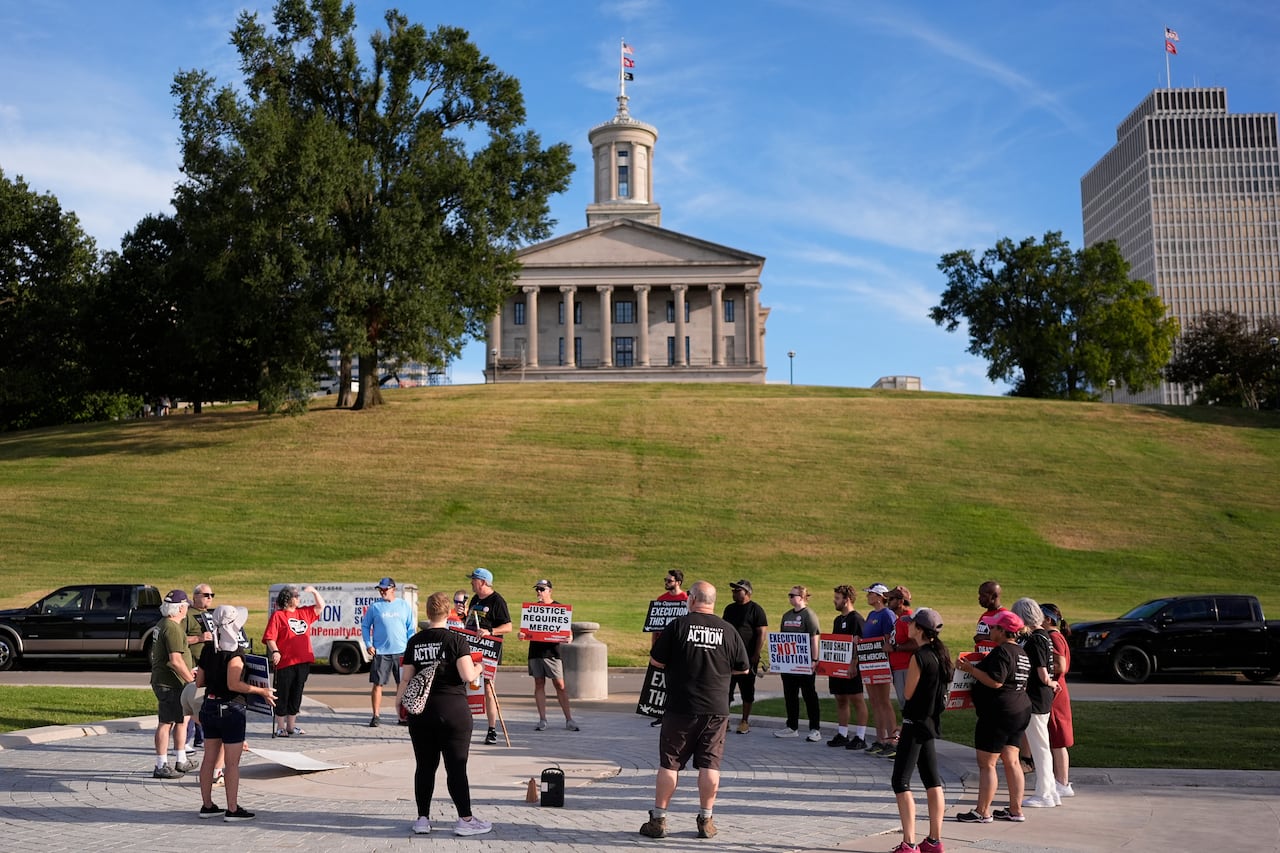 A group of people stand in a circle in front of a building atop a grassy hill.