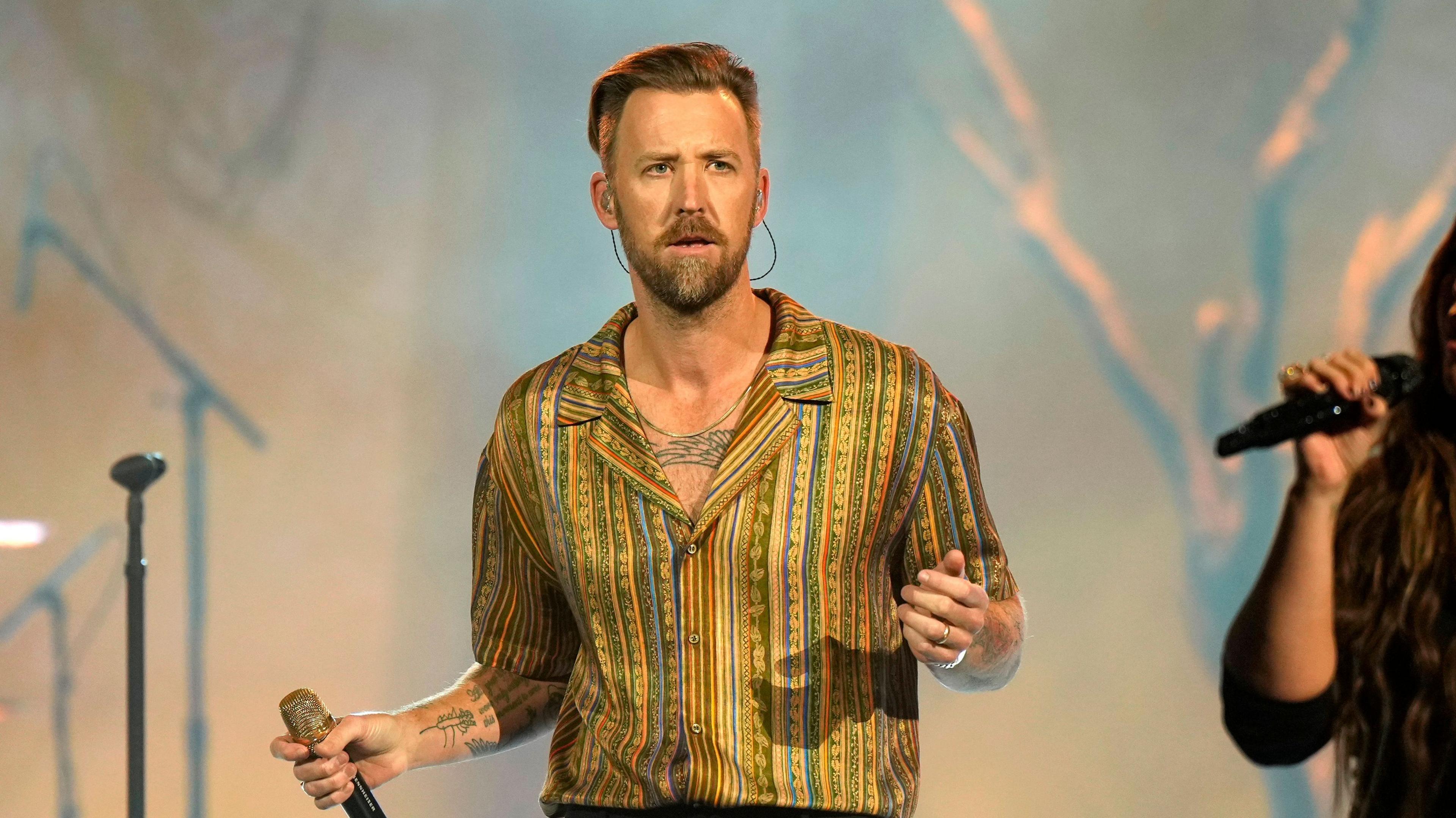 Charles Kelley of Lady A performing onstage during the 2024 People's Choice Country Awards at Grand Ole Opry House in Nashville, Tennessee. He is wearing a gold stripey shirt and holding a microphone.