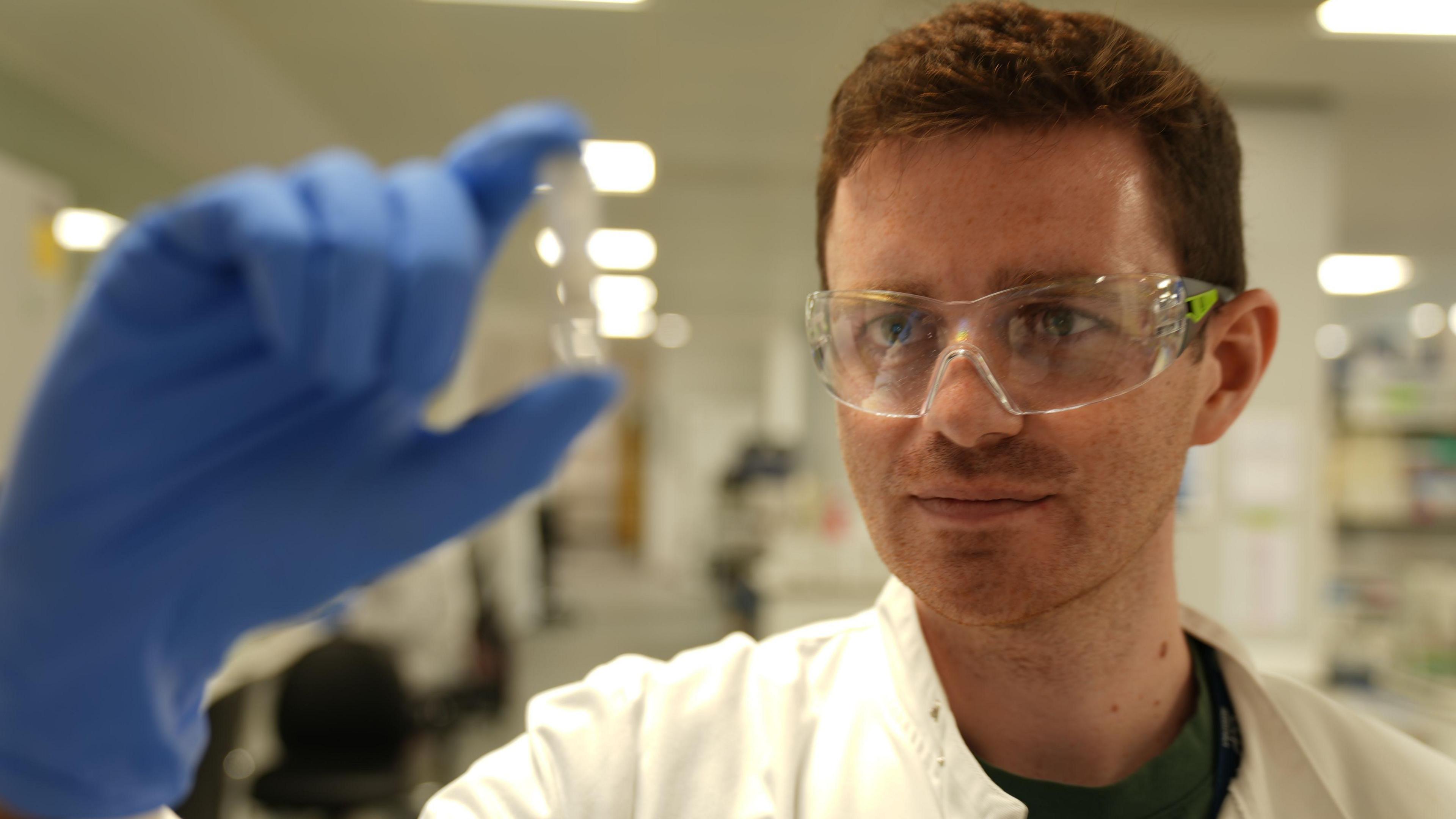 The image shows a man in a laboratory wearing a white lab coat, clear protective safety glasses, and a blue glove. The gloved hand is held up in the foreground, pinching a small transparent container or vial, which is slightly out of focus due to the shallow depth of field. The person is positioned on the right side of the frame and appears to be closely examining the object being held up. The background contains bright overhead lights, laboratory furnishings, and equipment, all blurred to empha