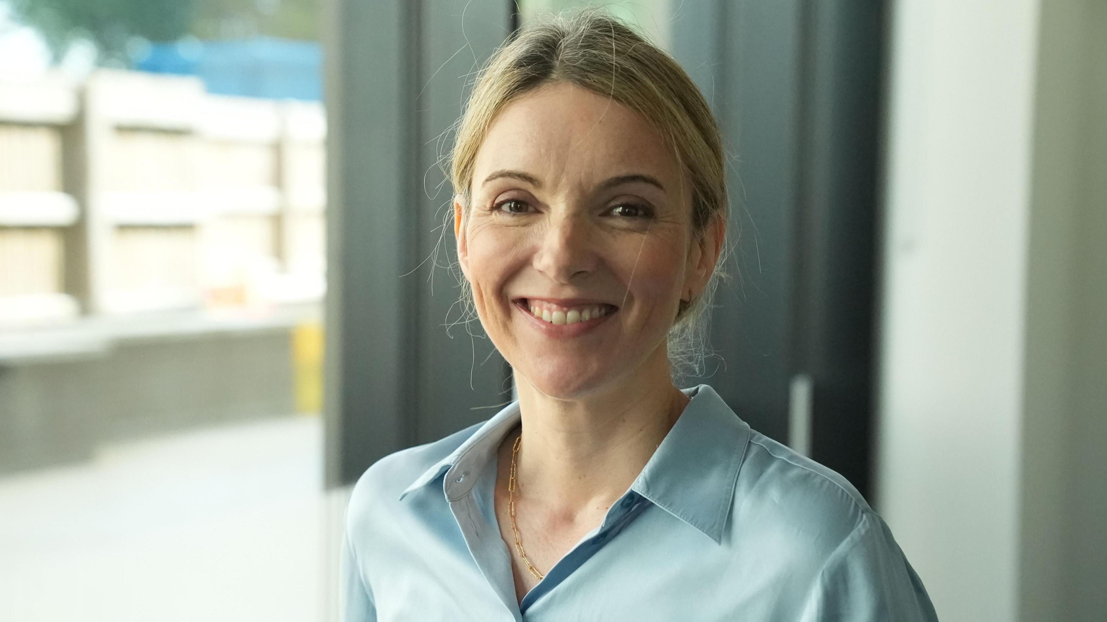 The image shows a person standing indoors near large windows in a bright, modern setting. The person is wearing a light blue button-up shirt and a thin necklace, with hair pulled back away from the face. The photograph is framed as a head-and-shoulders portrait, with the subject positioned prominently in the foreground. The background is softly blurred and includes window frames, a curtain, and an outdoor area visible through the glass. Natural light illuminates the scene, creating a clean and p