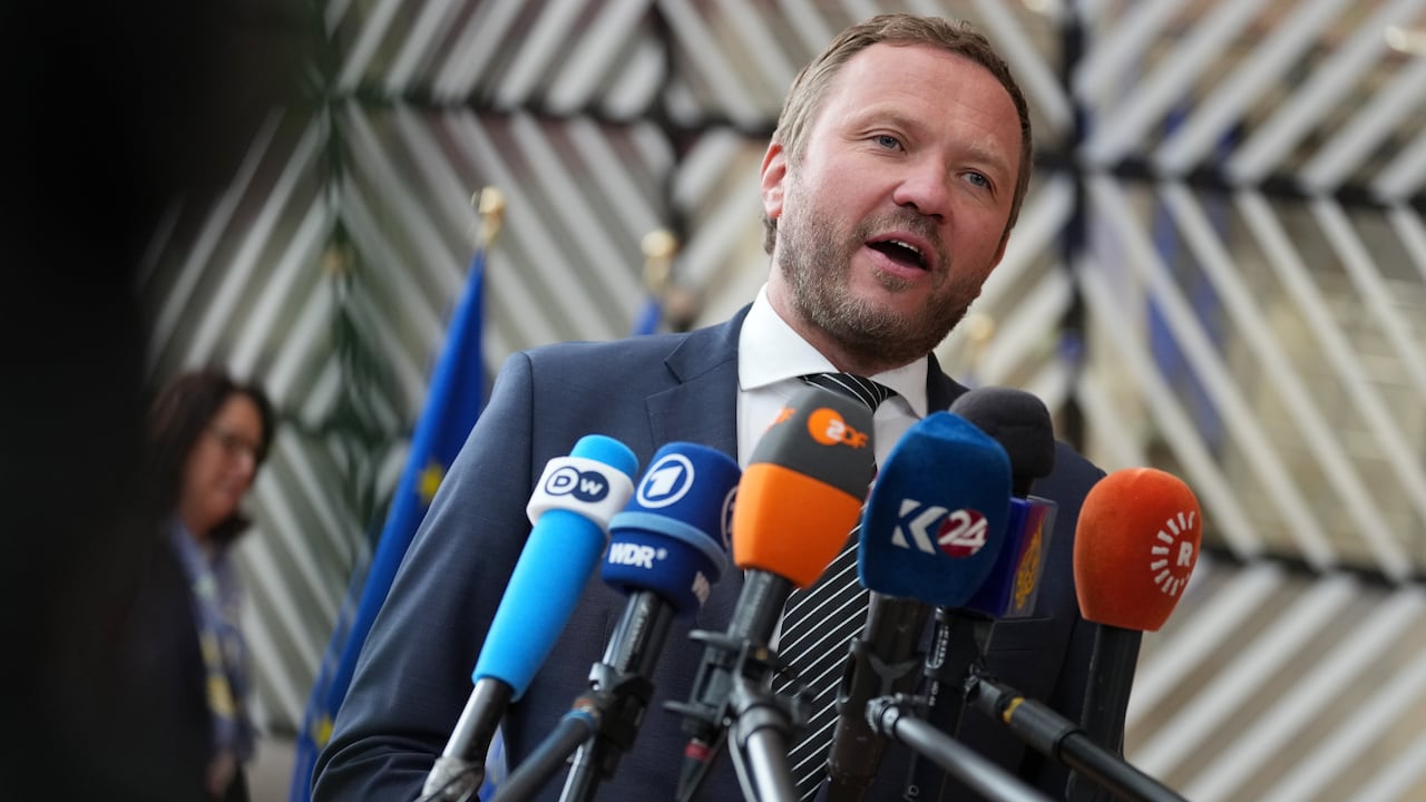 A bearded man with short hair wearing a suit and tie spreaks into a bank of several microphones.