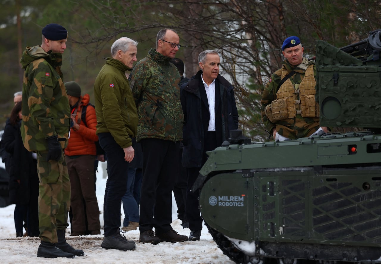 A handful of men in civilian clothing and a few others in military uniform examine a small tank-like machine in an outdoor setting.