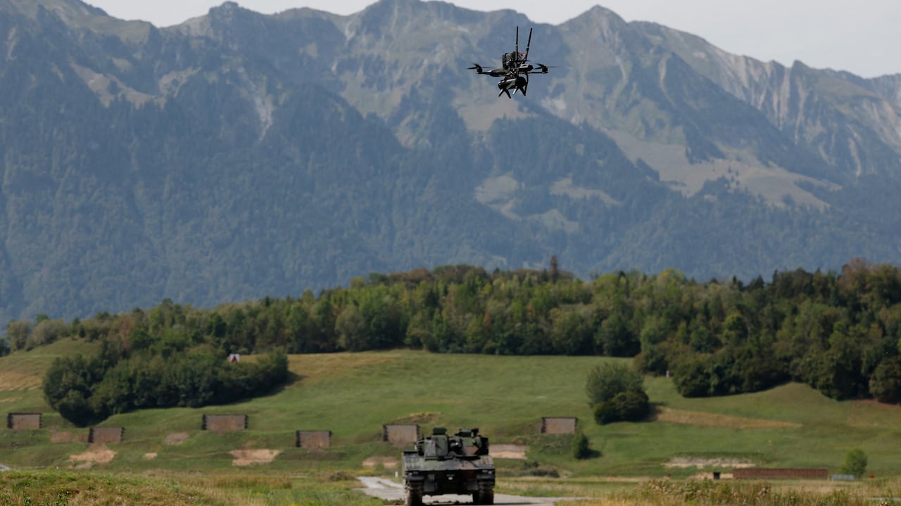 A drone is seen in the air with a tank on a road below it and a mountain range in the background.