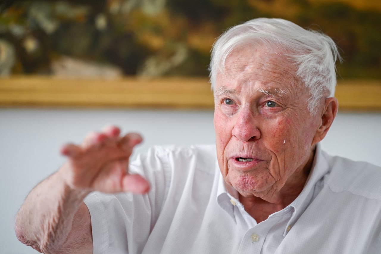 An old man in a white polo speaks at a table.