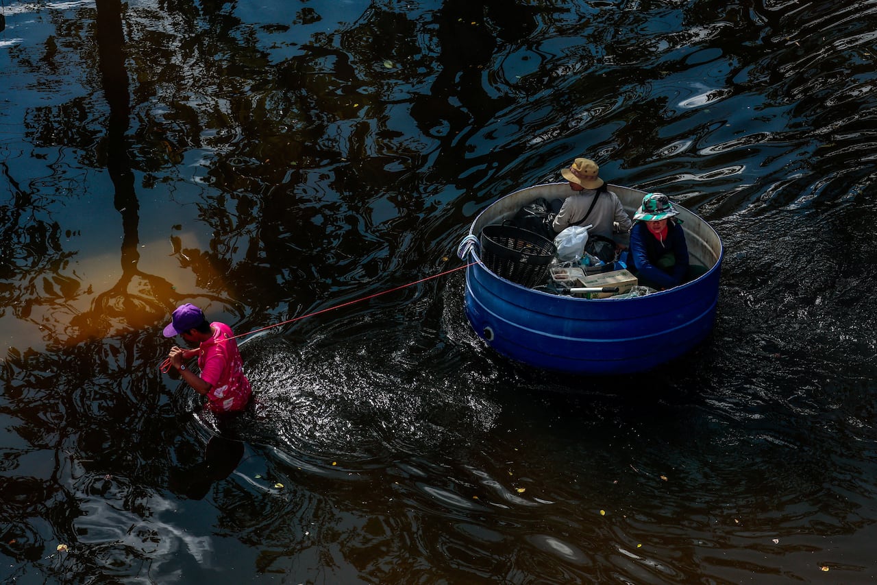 A man in hip-deep water pulls a red cord over his shoulder attached to a circular blue basin with two people in it.