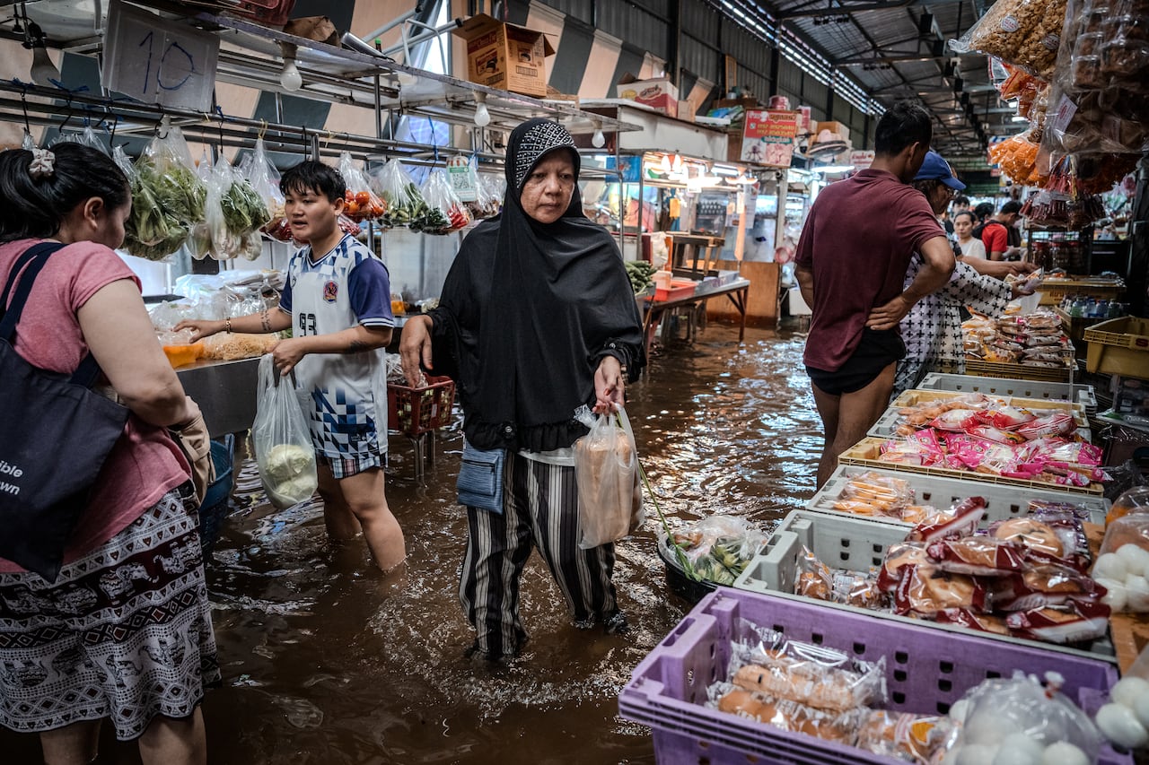 A woman in a headscarf with water halfway up her shins holds a bag and looks at wares in a market.