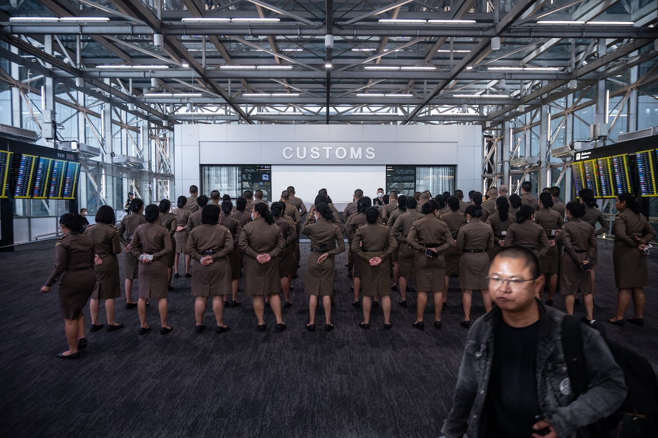 A man walks past women standing in rows under a sign that says "CUSTOMS".