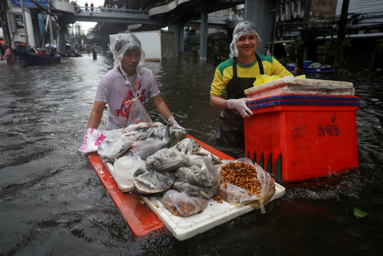 Two men walk through hip-deep water with plastic on the tops of their heads. One carries a large orange container, the other pushes items in bags on top of two stacked floating platforms, one orange and one white.
