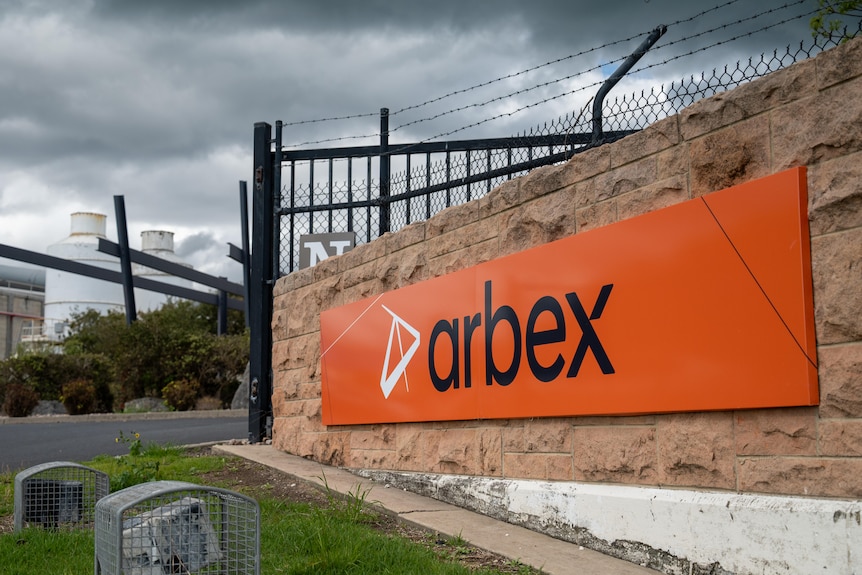 an orange sign on a small wall in front of a factory, reading Arbex