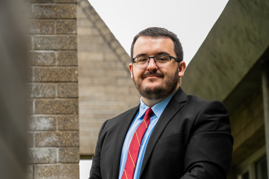 Jason Virgo, in glasses and a suit, standing next to a brick wall.