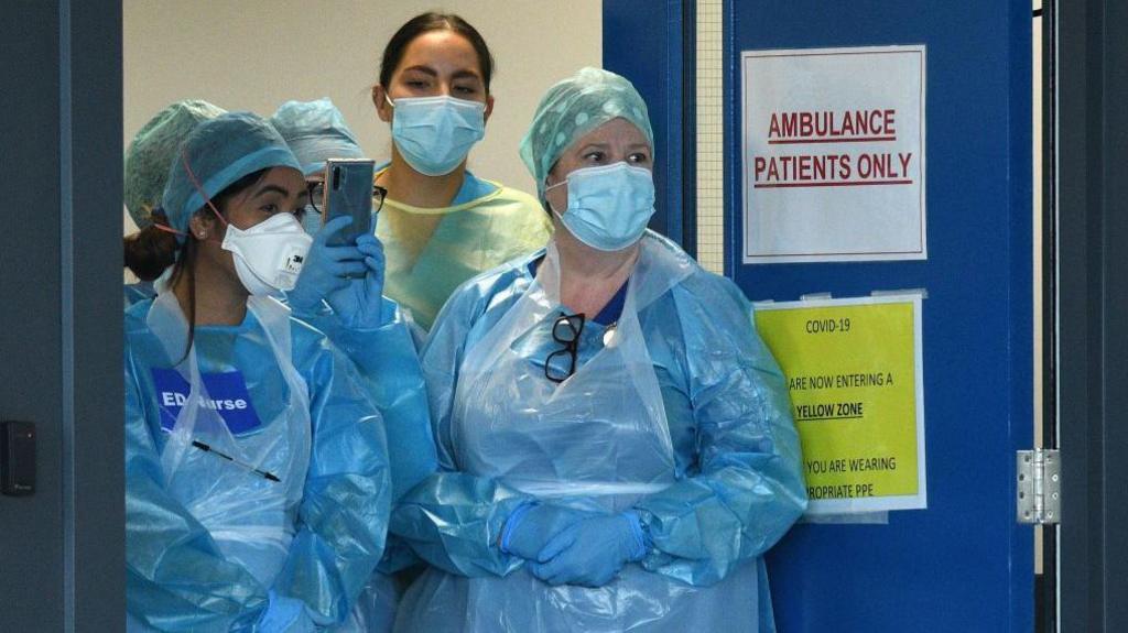 Four medics are standing at the doors of an emergency department in the pandemic. They are wearing plastic blue gowns and have face masks on. One has a sign on her chest saying "ED Nurse". 