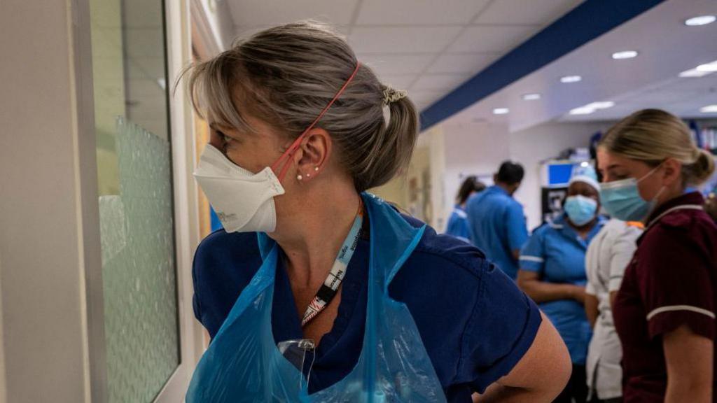 A medic standing in what looks like the emergency room of an NHS hospital in Covid. She is staring through a window perhaps looking outside. She is wearing a plastic blue apron over her scrubs and has a white masks on. Behind her are other members of staff wearing PPE. 
