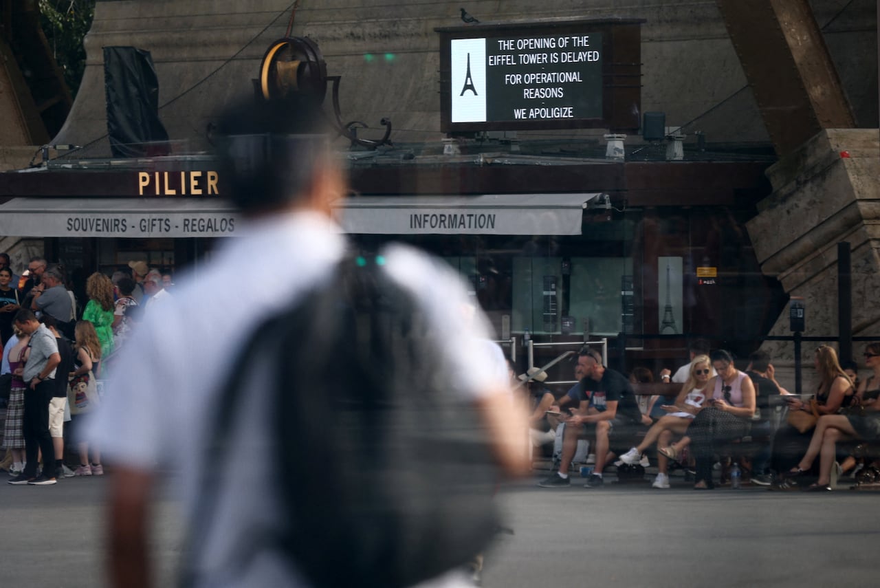 A man with a backpack walks near the Eiffel Tower, which has a sign reading the its opening is delayed for operational reasons.