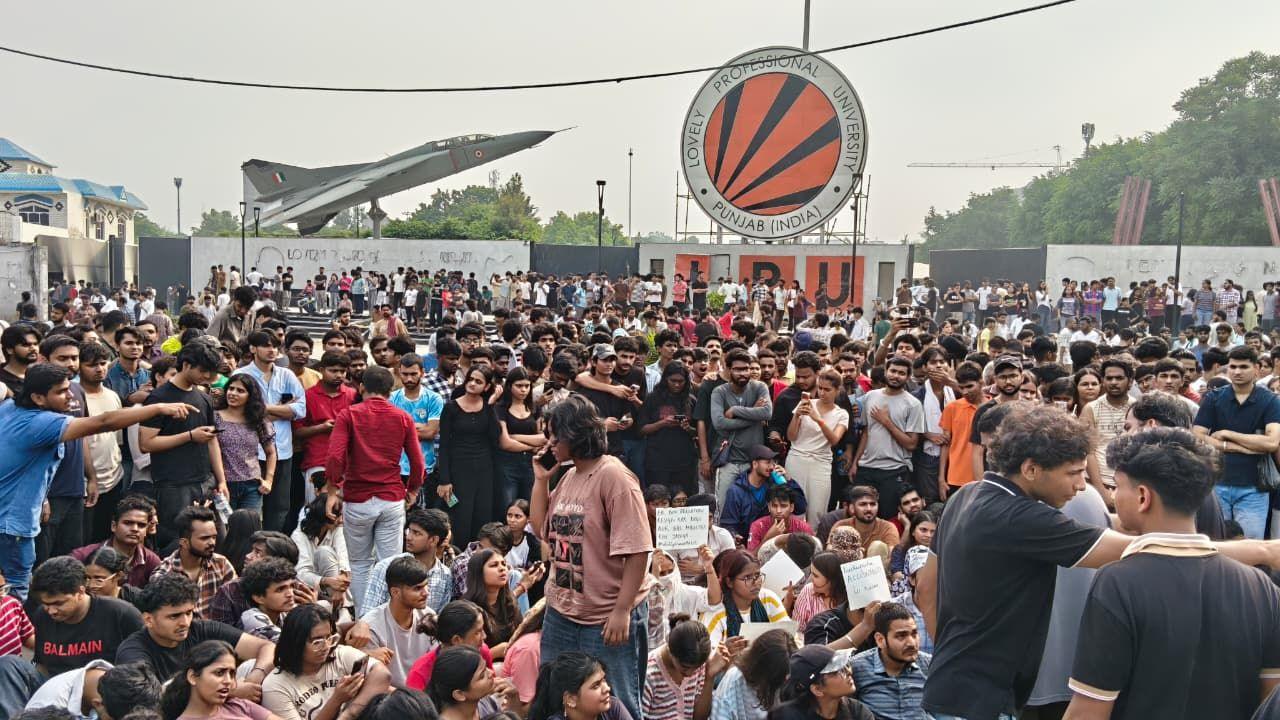 Students sitting on the ground and standing and an IAF aircraft and Lovely Professional University logo seen in the background. 