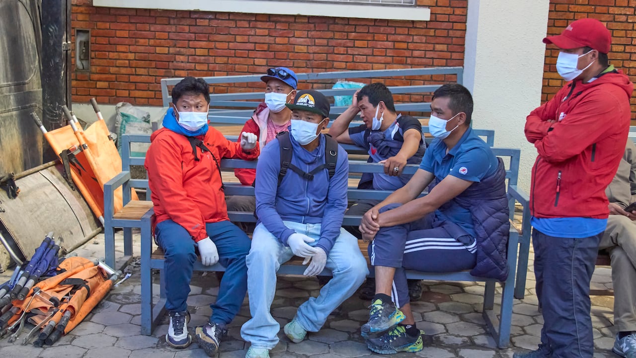 A group of men wearing athletic clothing and face masks sit on benches and stand outside a building.