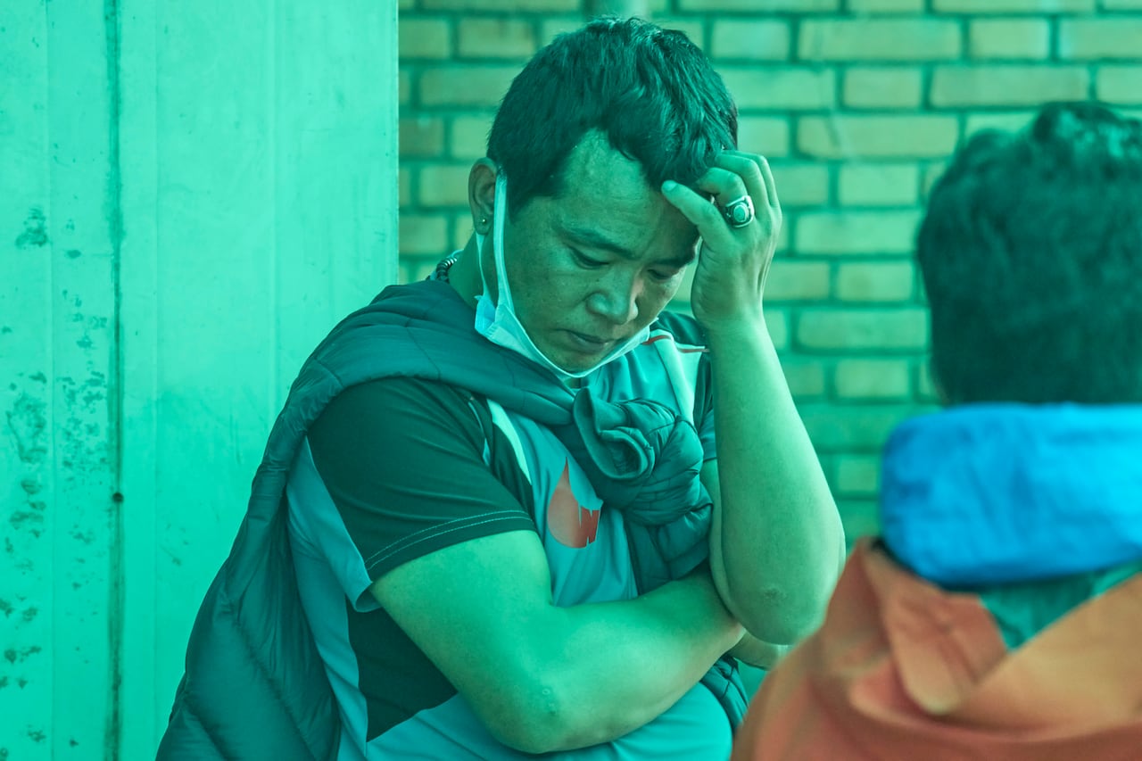 A man, with a face mask pulled down below his chin, stands with his head bowed and a sombre expression on his face.