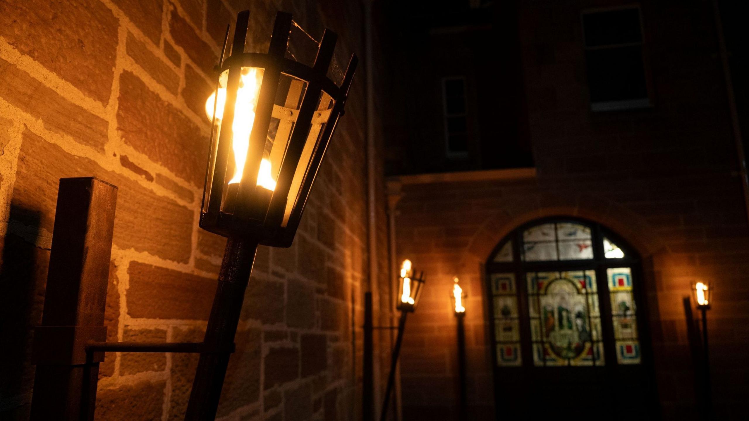 Burning torches fixed to stone walls inside Ardross Castle. The space is deep in shadow and there is a stained glass window on the far wall.